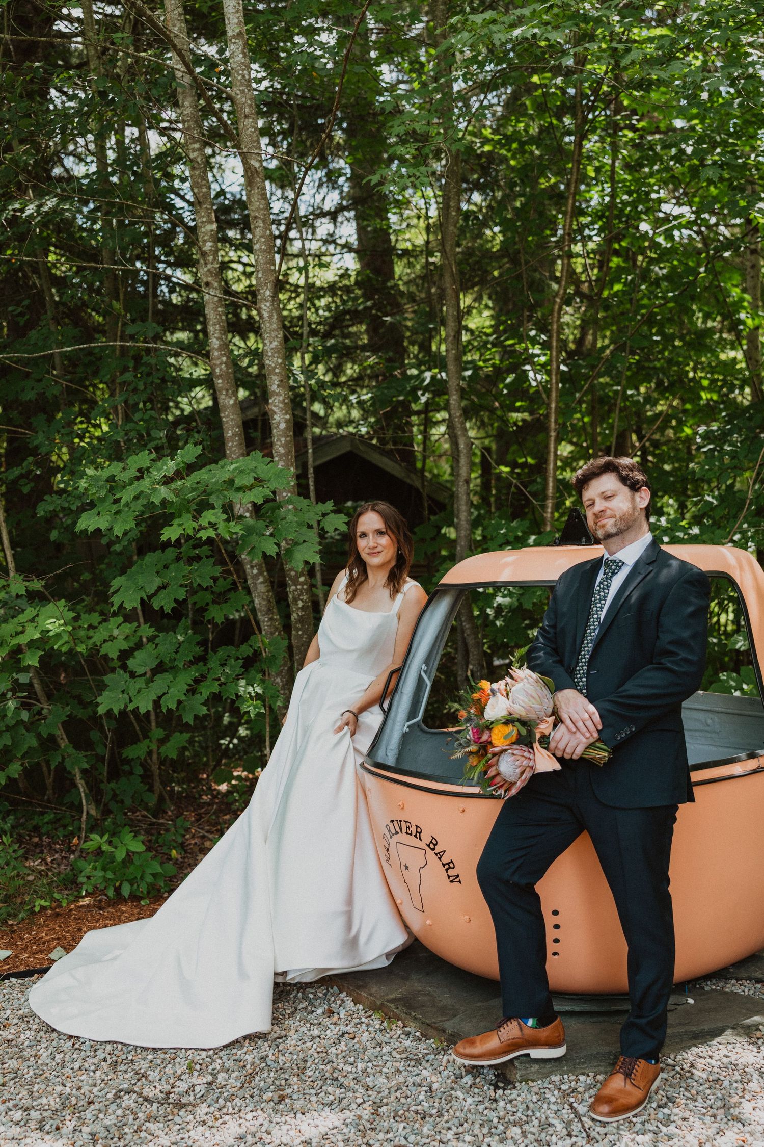 Bride and groom posing beside vintage orange car in wooded setting on their wedding day.