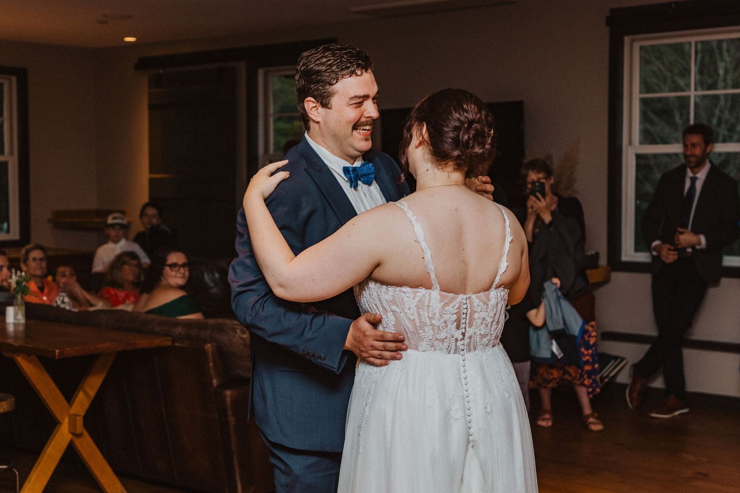 Smiling groom in navy suit shares first dance with bride in lace gown at indoor wedding reception.