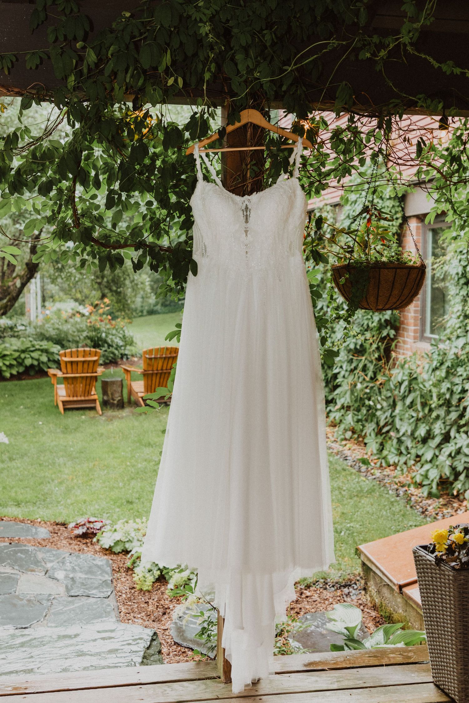 A white wedding dress hanging on a wooden hanger outdoors, surrounded by lush green garden foliage.