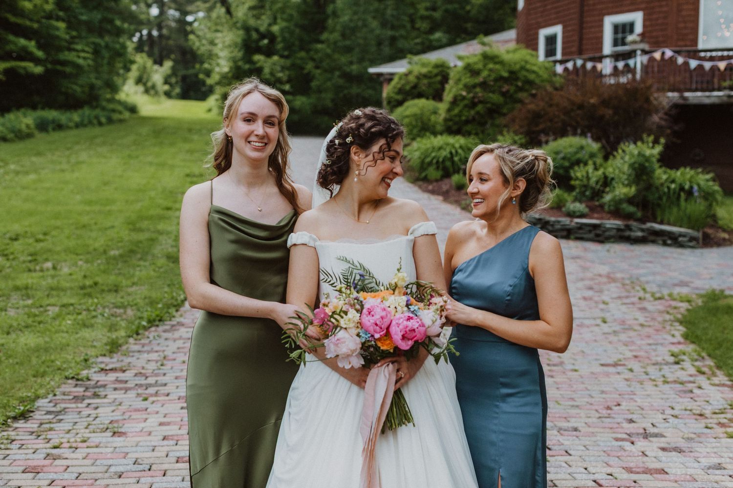 Bride in white off-shoulder gown walks with two bridesmaids in olive and blue dresses, holding colorful bouquet outdoors.