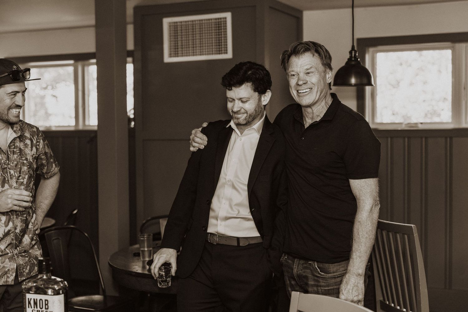 Two men smiling and embracing at an indoor gathering, one in a blazer and the other in a black shirt.
