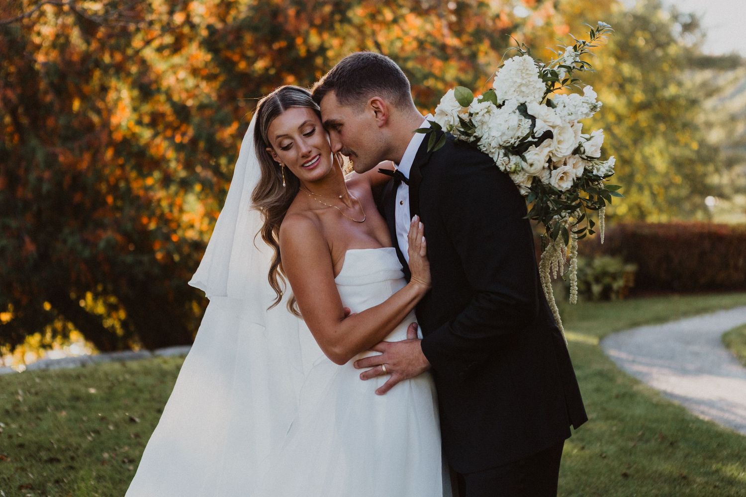 Groom in black tuxedo tenderly kisses bride in white gown amid vibrant fall foliage backdrop.