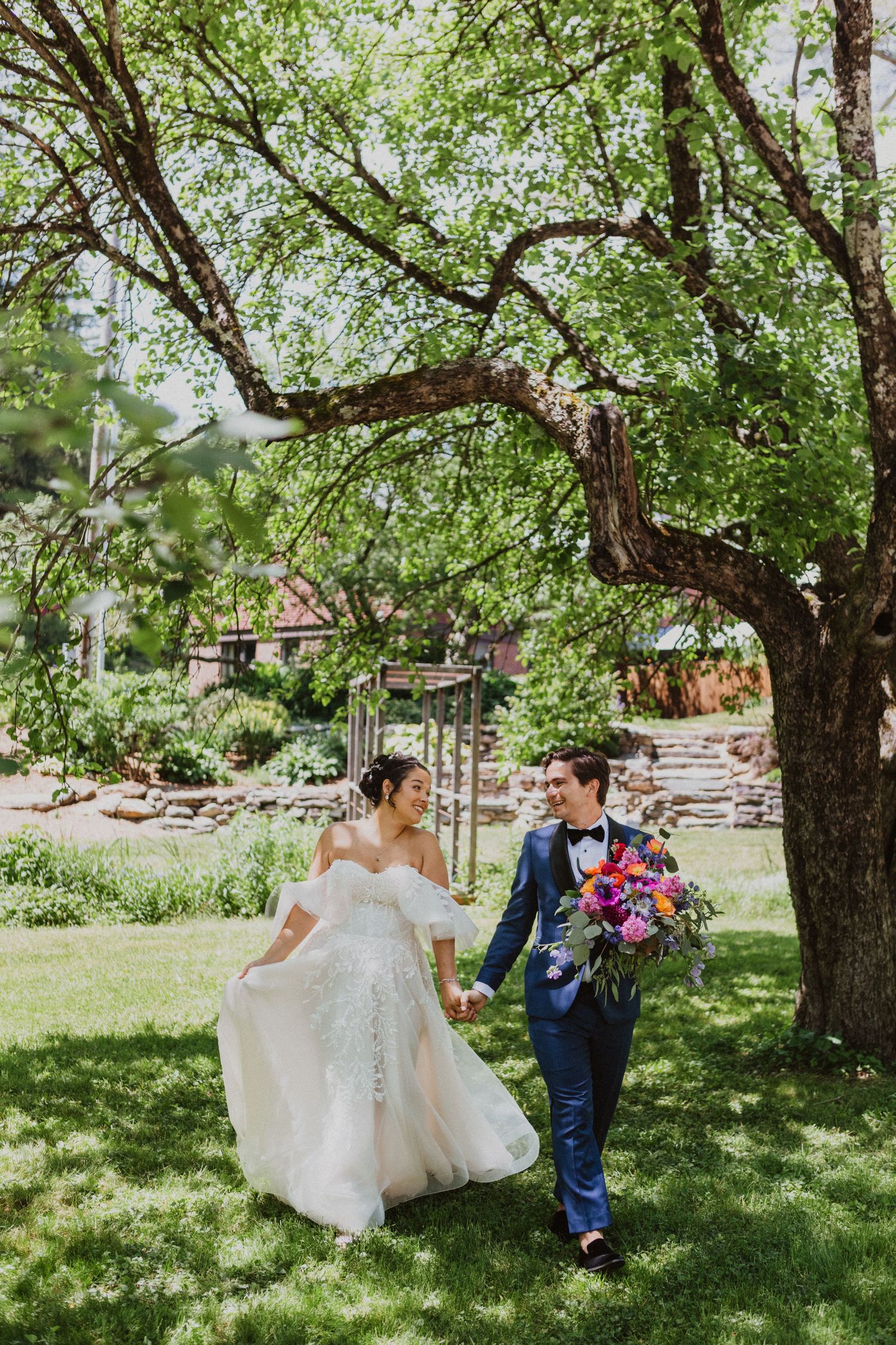 A bride in a lace gown and groom in a navy suit walk hand in hand beneath a large tree in a garden.