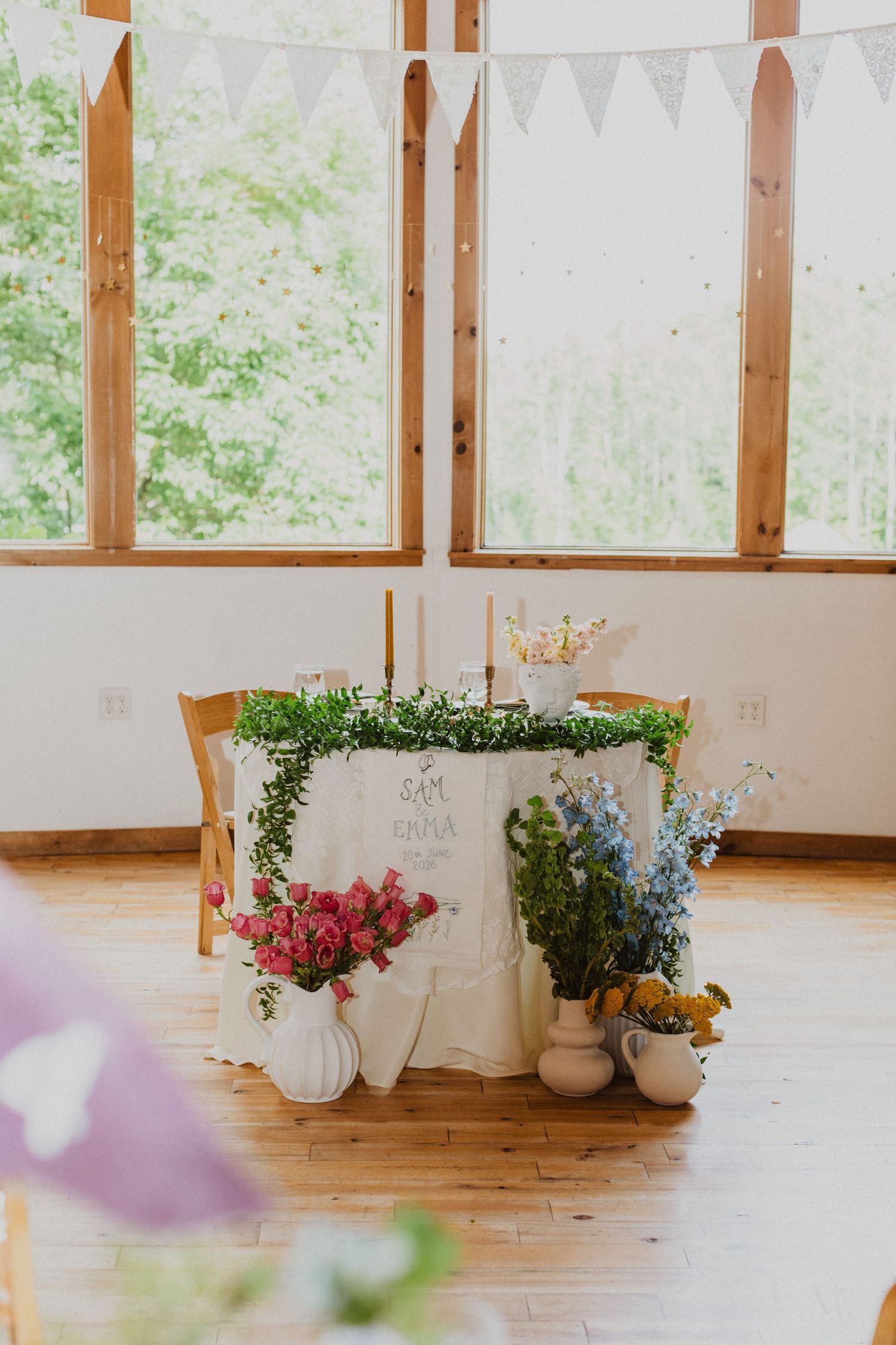 Elegant wedding sweetheart table with lush floral arrangements, greenery garland, and bunting in a bright barn venue.