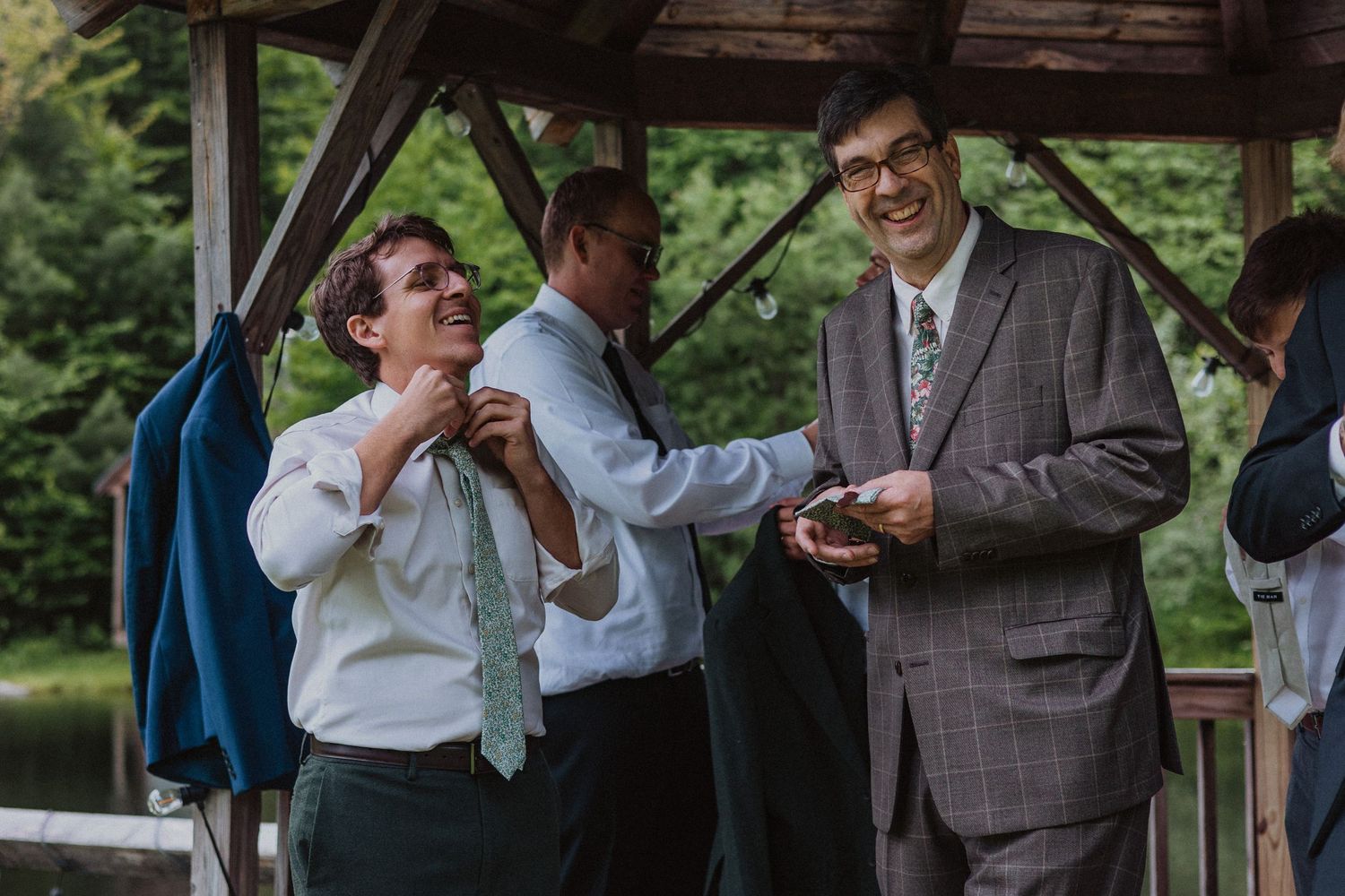 Men in formal attire laughing and adjusting ties at an outdoor wedding venue with lush greenery.