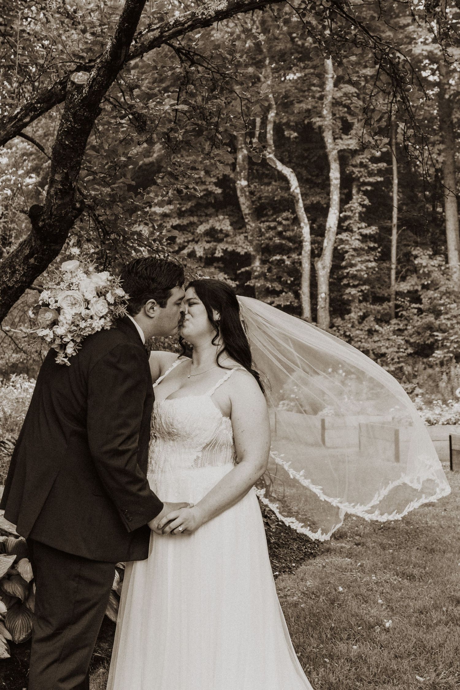 Bride and groom share a romantic kiss outdoors with veil flowing in the wind, surrounded by lush green trees.