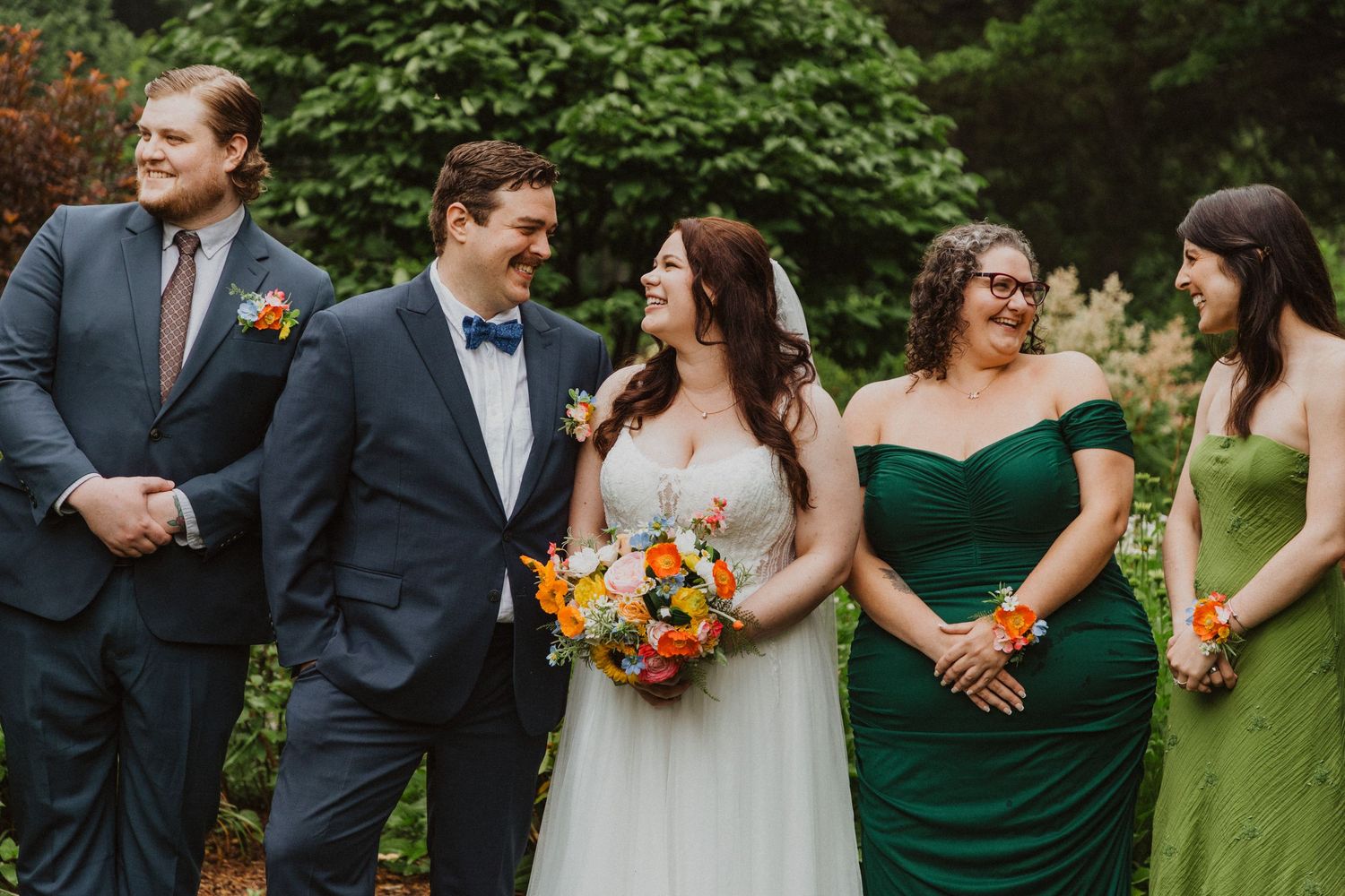 Wedding party laughs together outdoors, bridesmaids in green dresses with groomsmen in navy and blue suits.