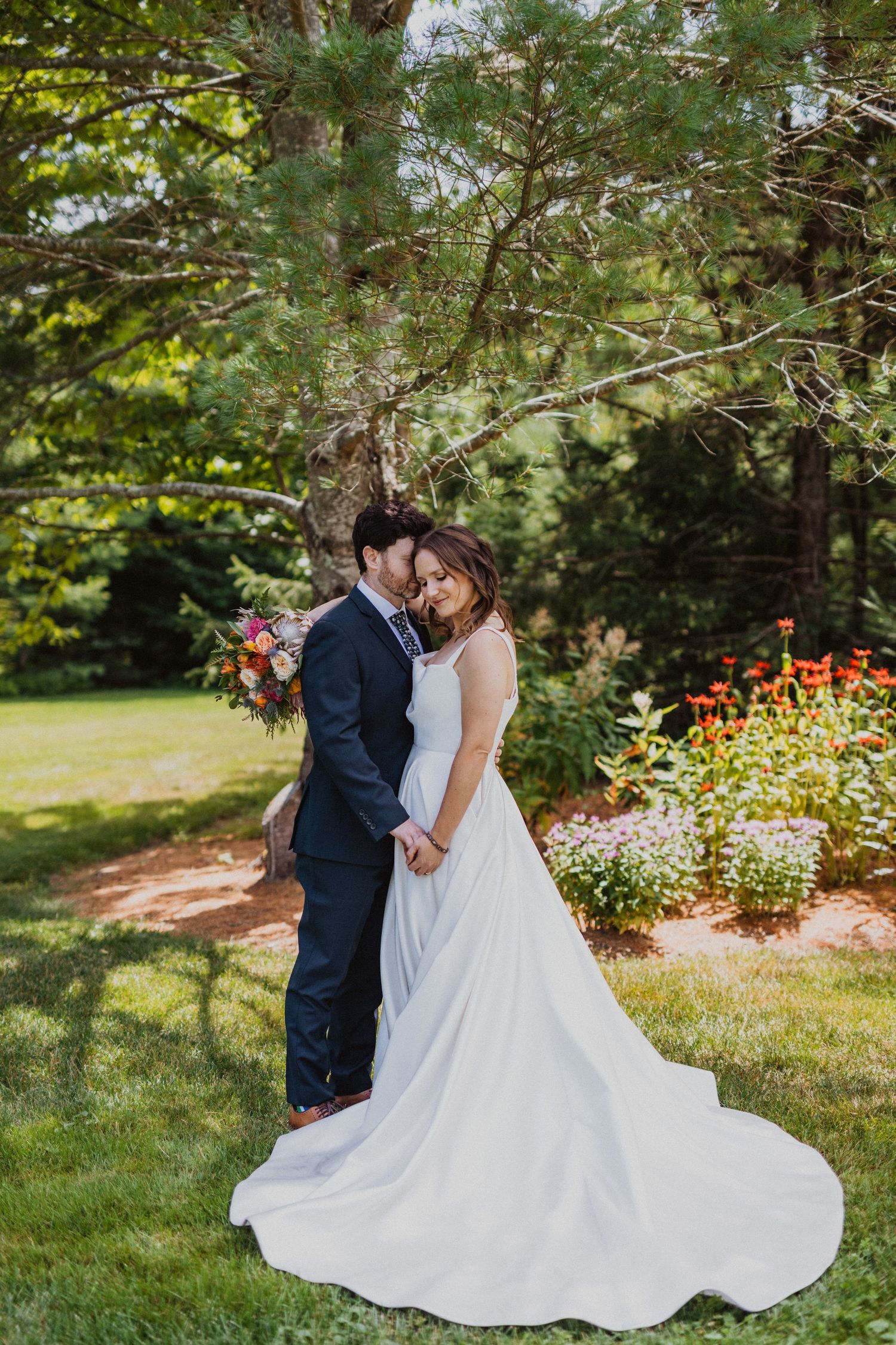 Bride and groom share a romantic embrace under a pine tree in a lush garden setting on their wedding day.
