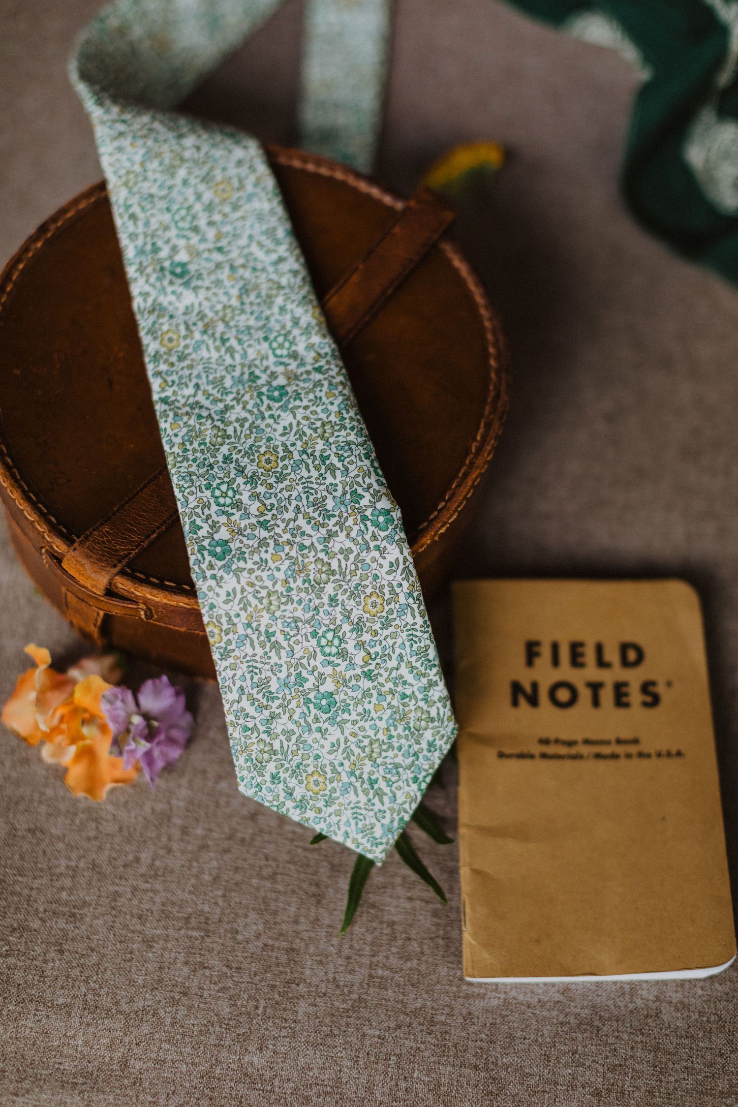 Floral green tie resting on a brown leather box beside a Field Notes notebook and colorful flowers.