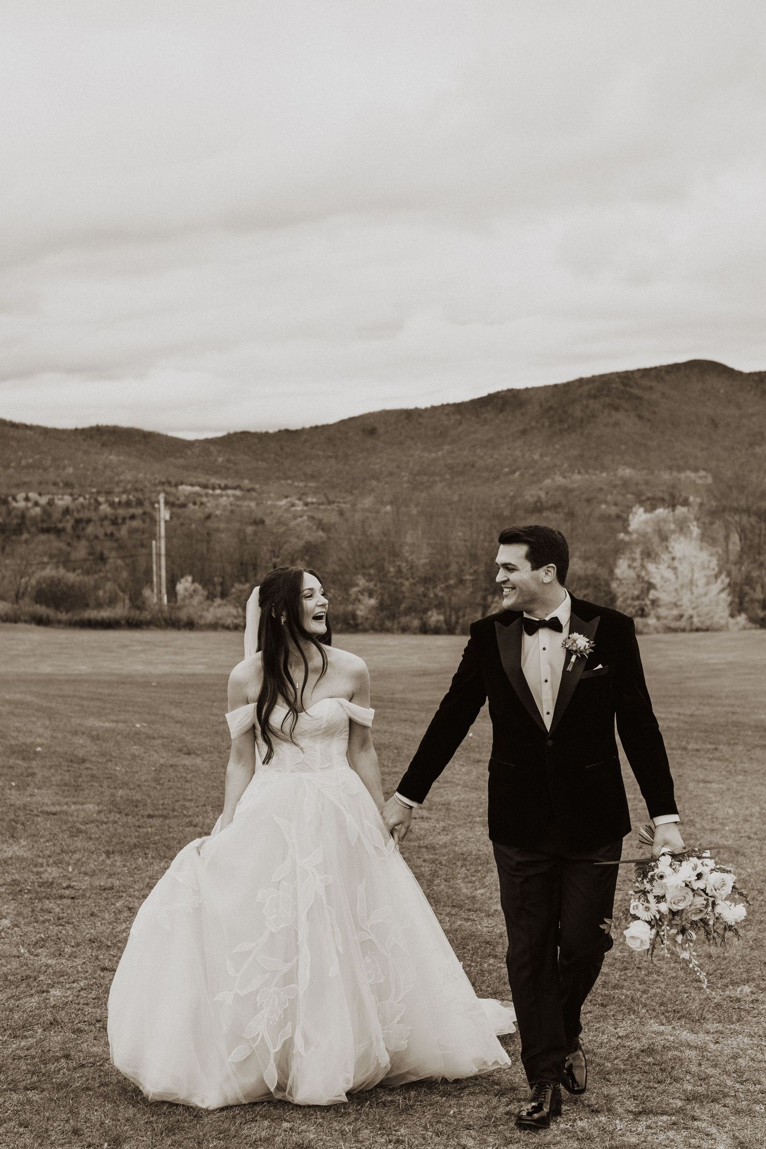 Black and white photo of laughing bride and groom walking hand in hand across mountain meadow.