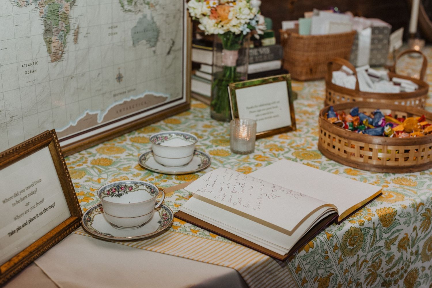 Wedding guest book table with vintage map, teacups, floral tablecloth, and candy basket display.