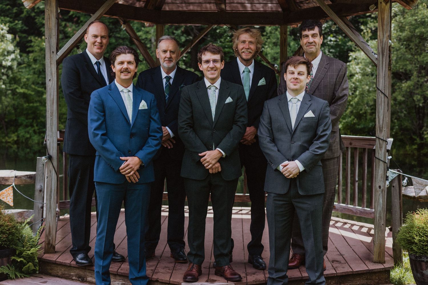 Seven men in suits pose together smiling under a wooden gazebo surrounded by lush green trees at a wedding.