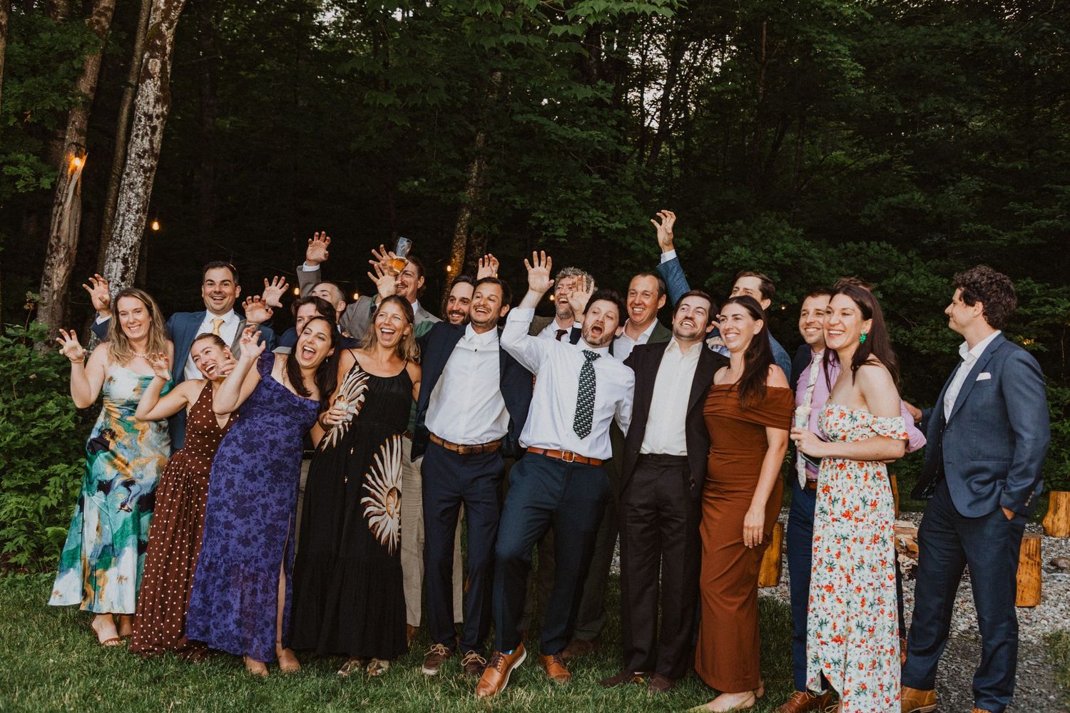 A joyful group of wedding guests celebrating outdoors, raising hands and cheering near a lush green forest backdrop at dusk.