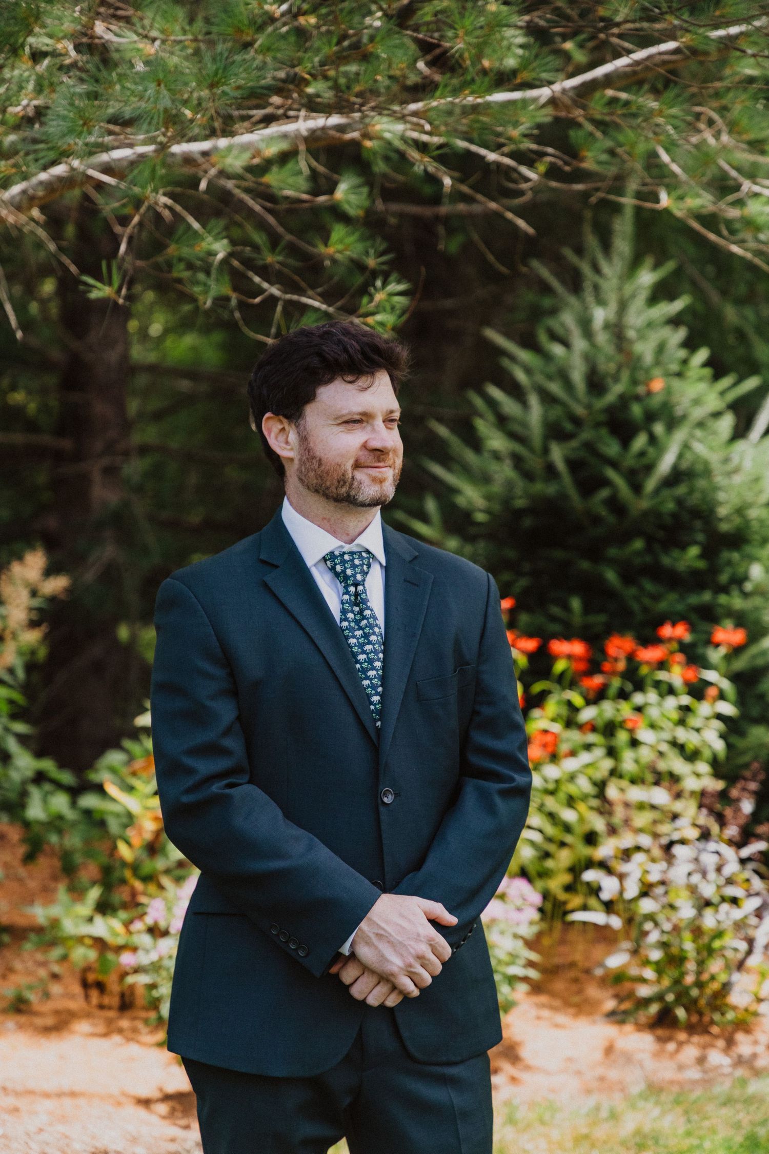 Groom in dark navy suit and floral tie standing in garden surrounded by pine trees and colorful flowers.