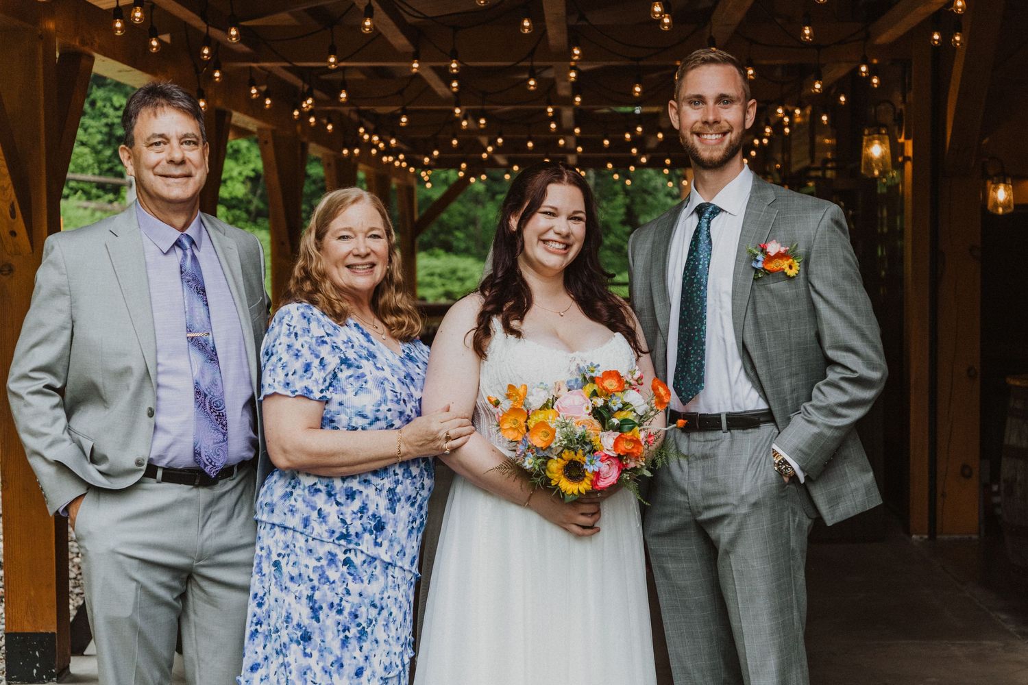 Bride with bouquet poses with family members at rustic wedding venue with string lights.