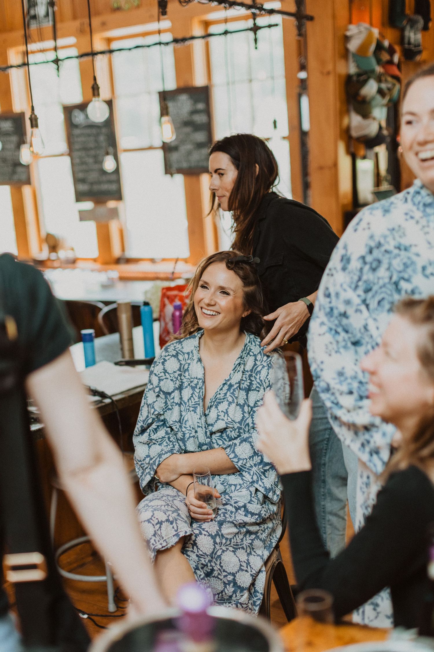 Bride smiling in blue floral robe getting hair done by stylist in rustic wood-paneled venue.