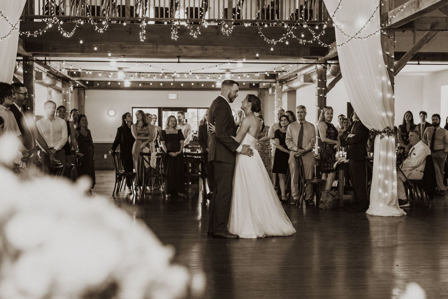 Bride and groom sharing first dance in elegant barn venue with string lights and guests.