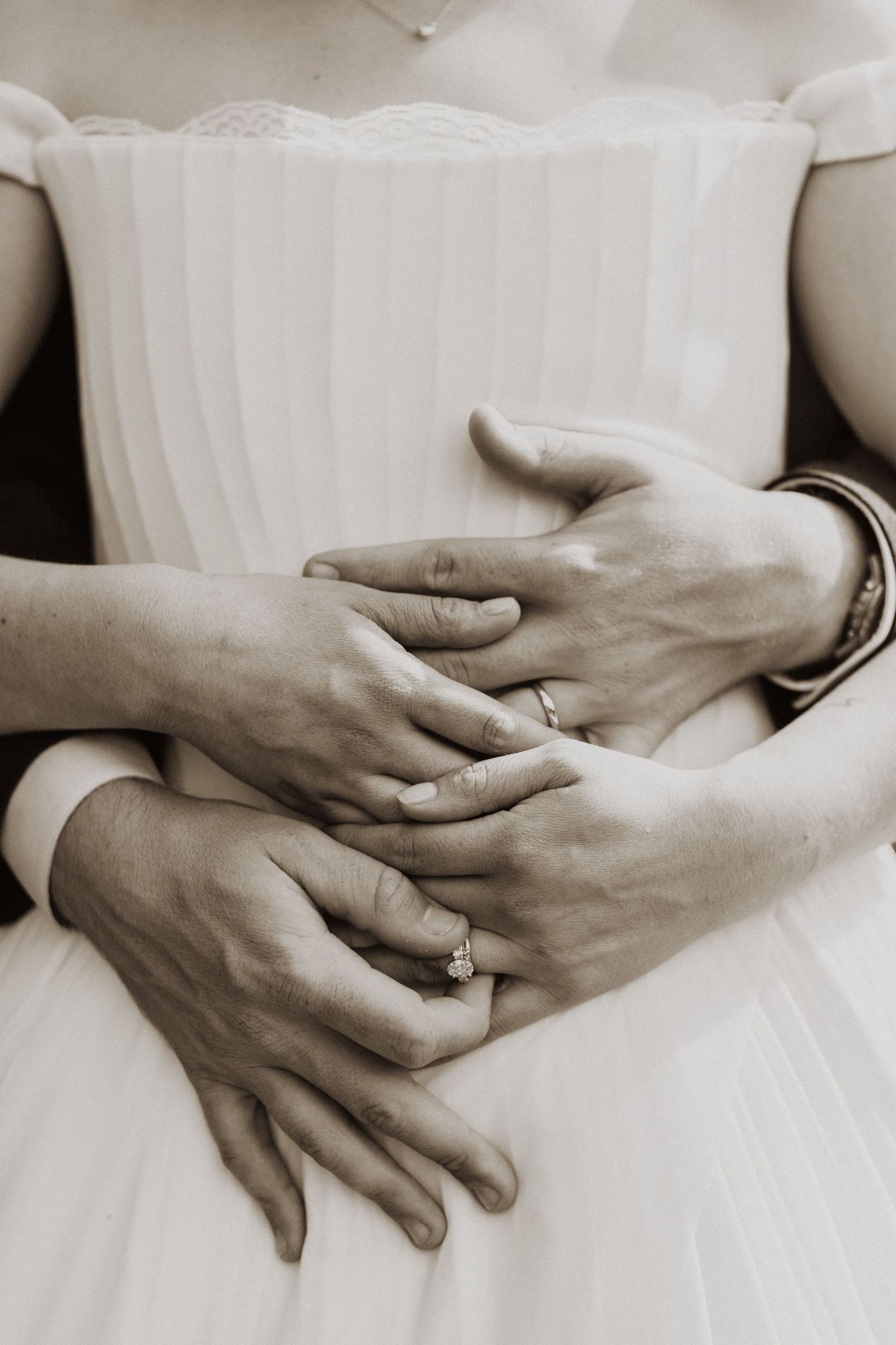 Close-up sepia photo of bride and groom hands embracing, showing diamond engagement ring and wedding bands.