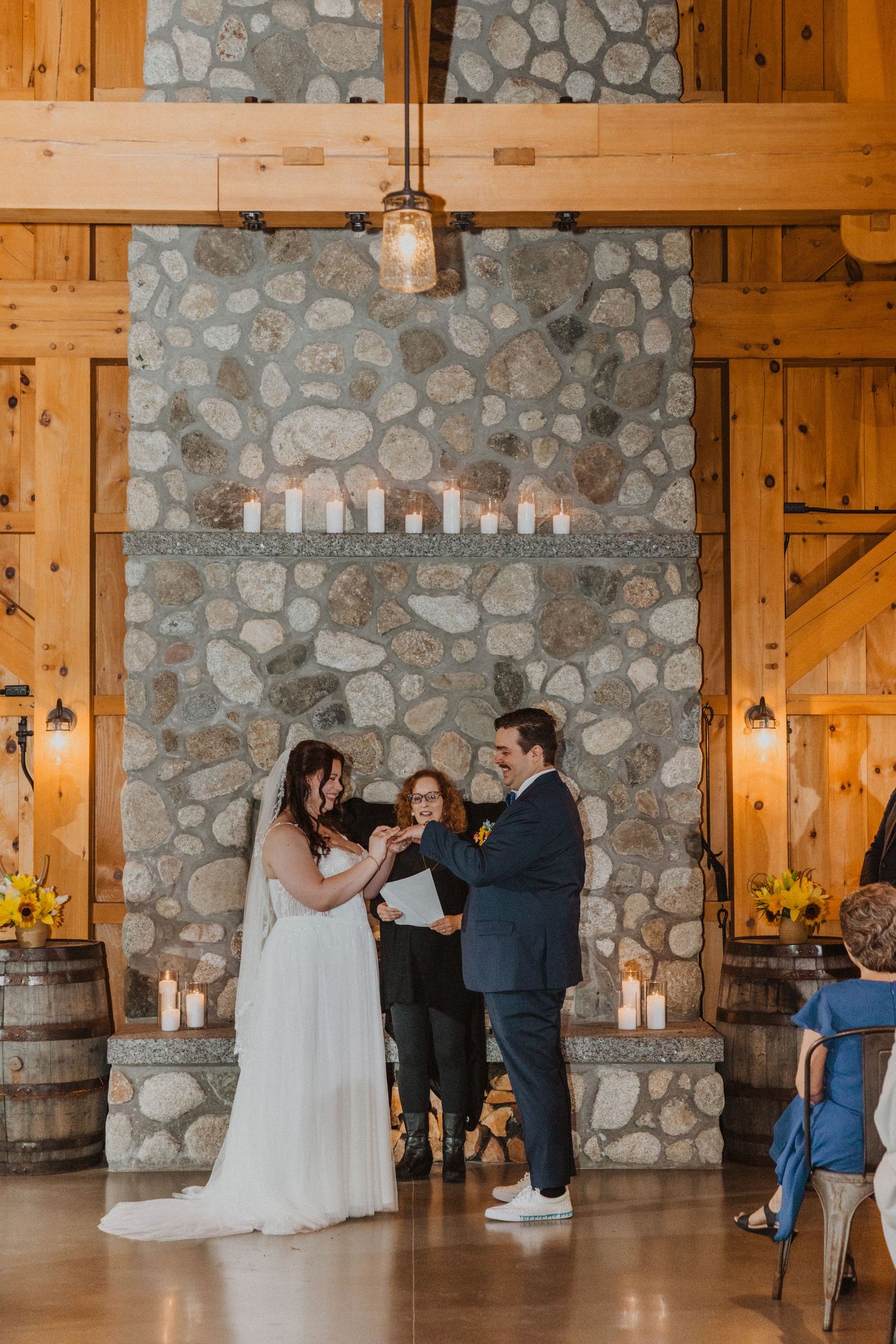 Couple exchanging rings at rustic barn wedding ceremony before stone fireplace with candles and wood beam decor.