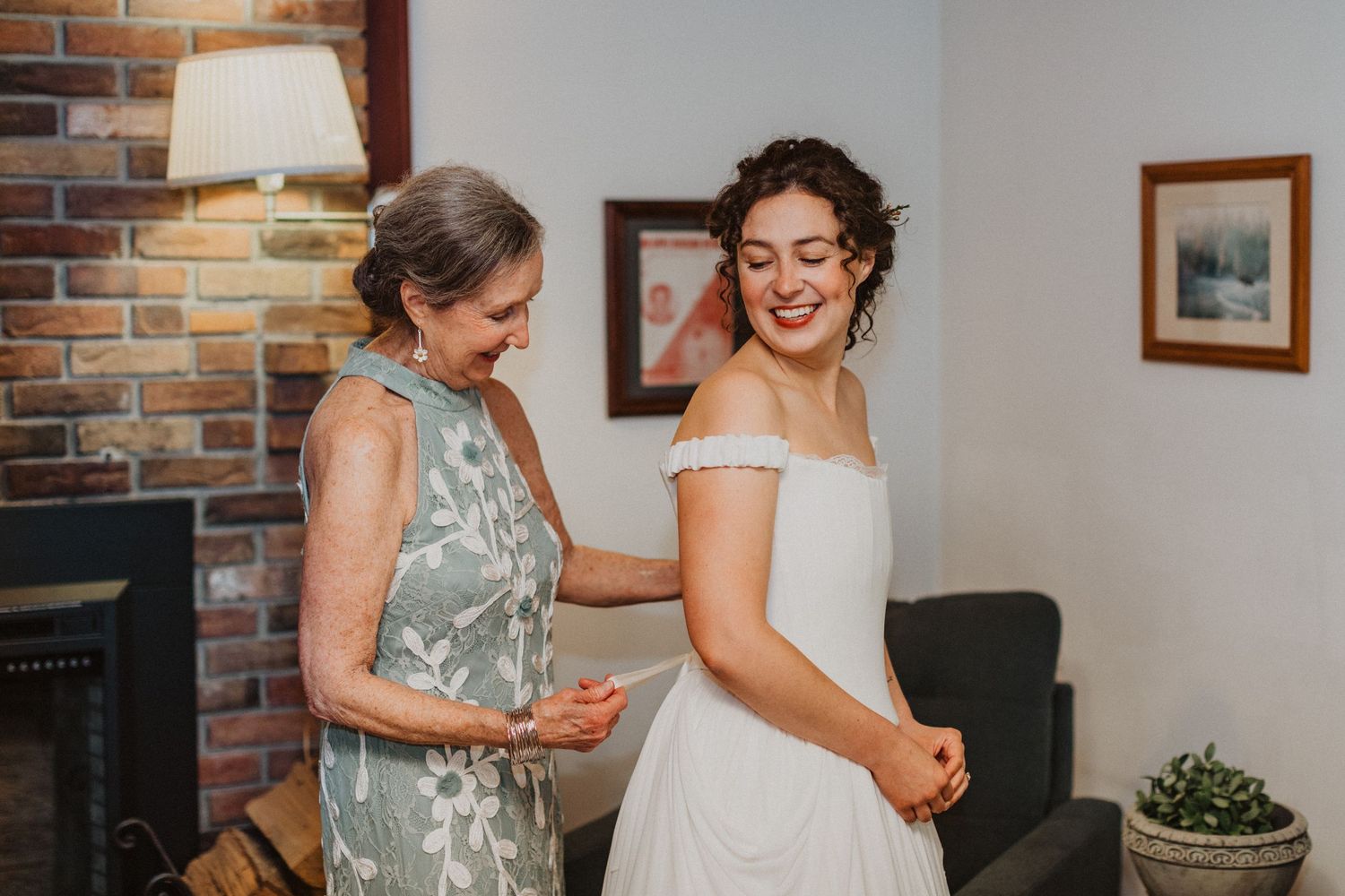Mother helping bride into off-shoulder white wedding dress in a cozy room with brick fireplace.