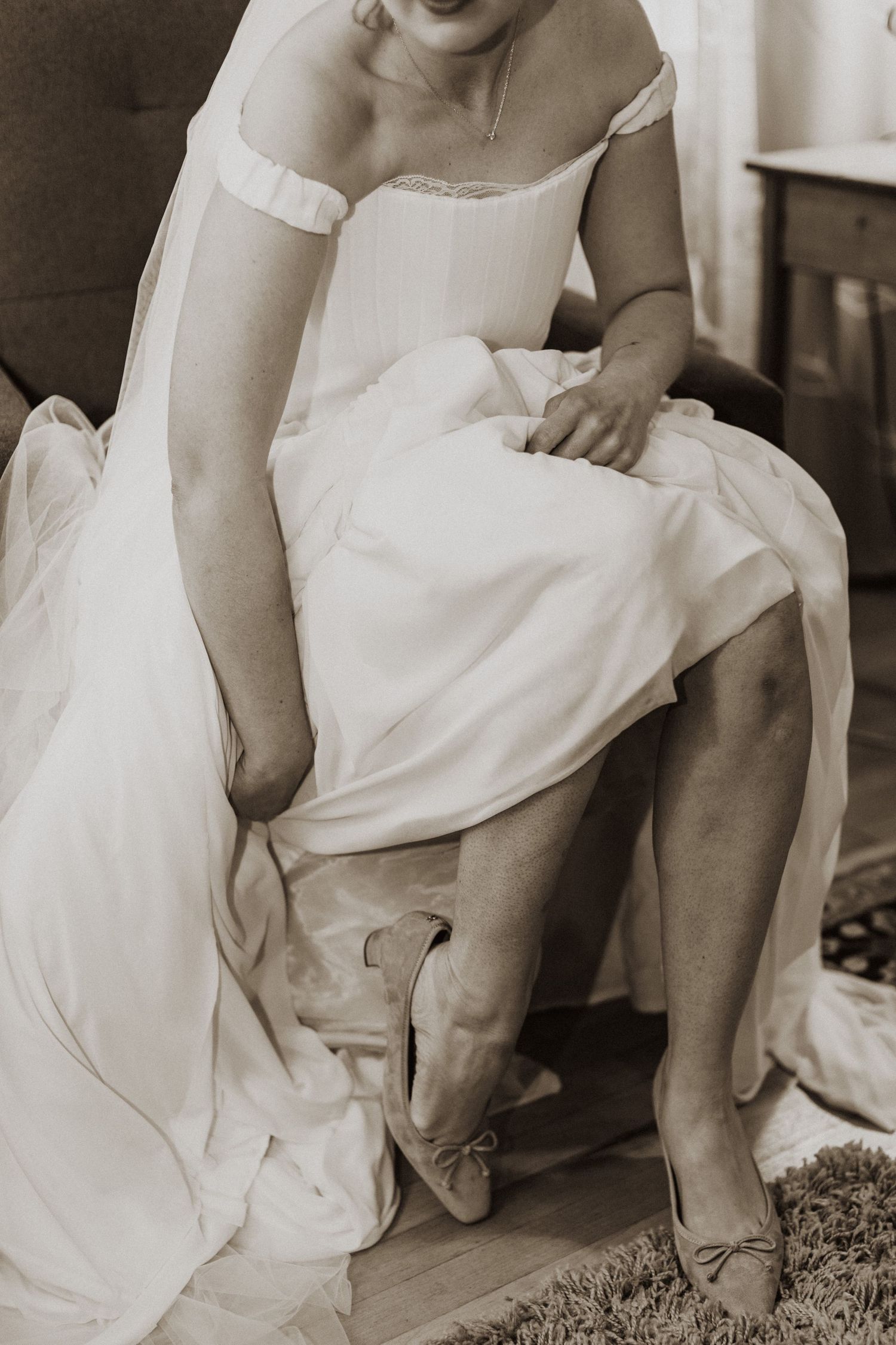 Bride in white off-shoulder gown adjusting bow heels while seated, sepia-toned wedding preparation photo.
