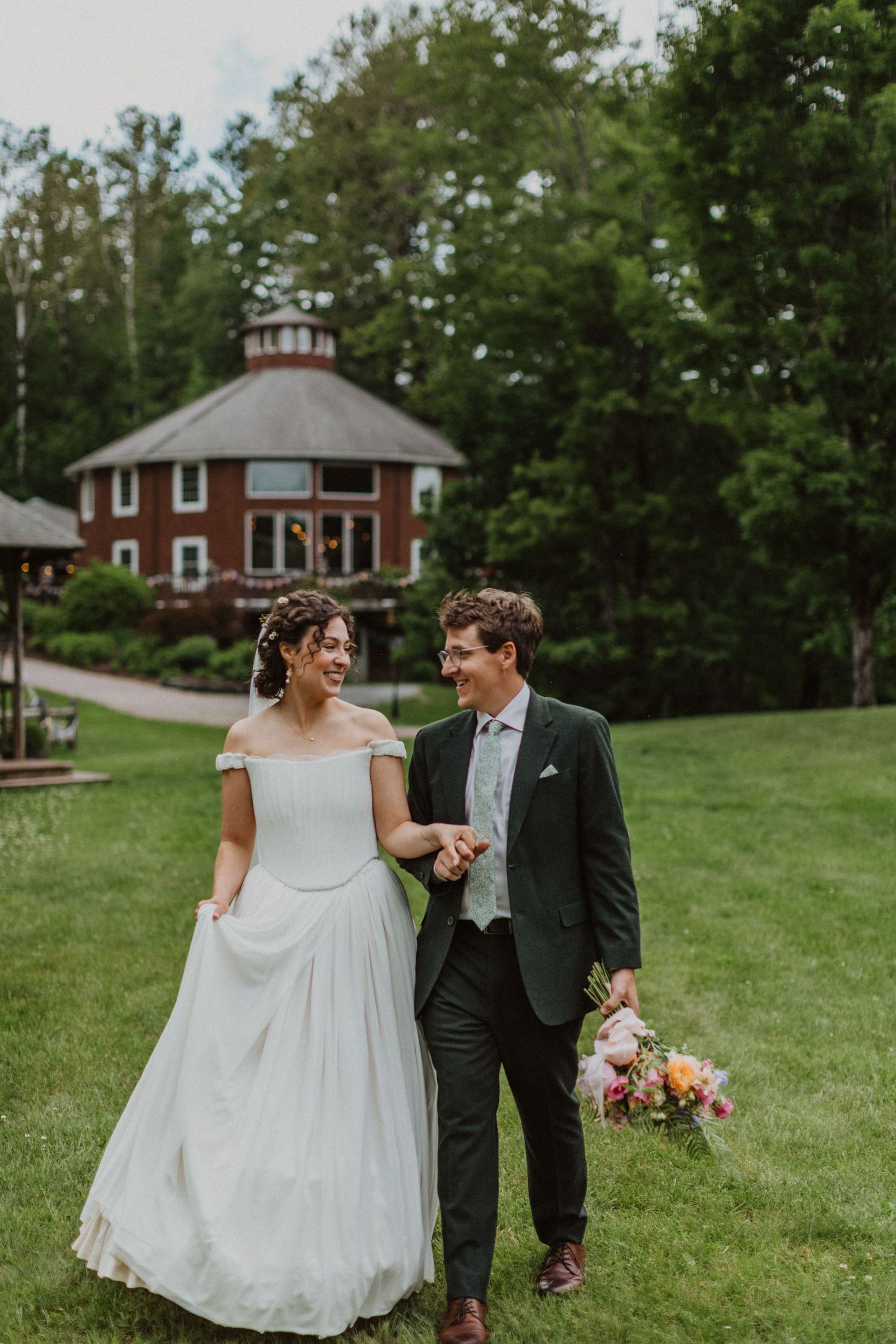 Smiling bride and groom walk hand-in-hand on green lawn with rustic red venue building behind them.