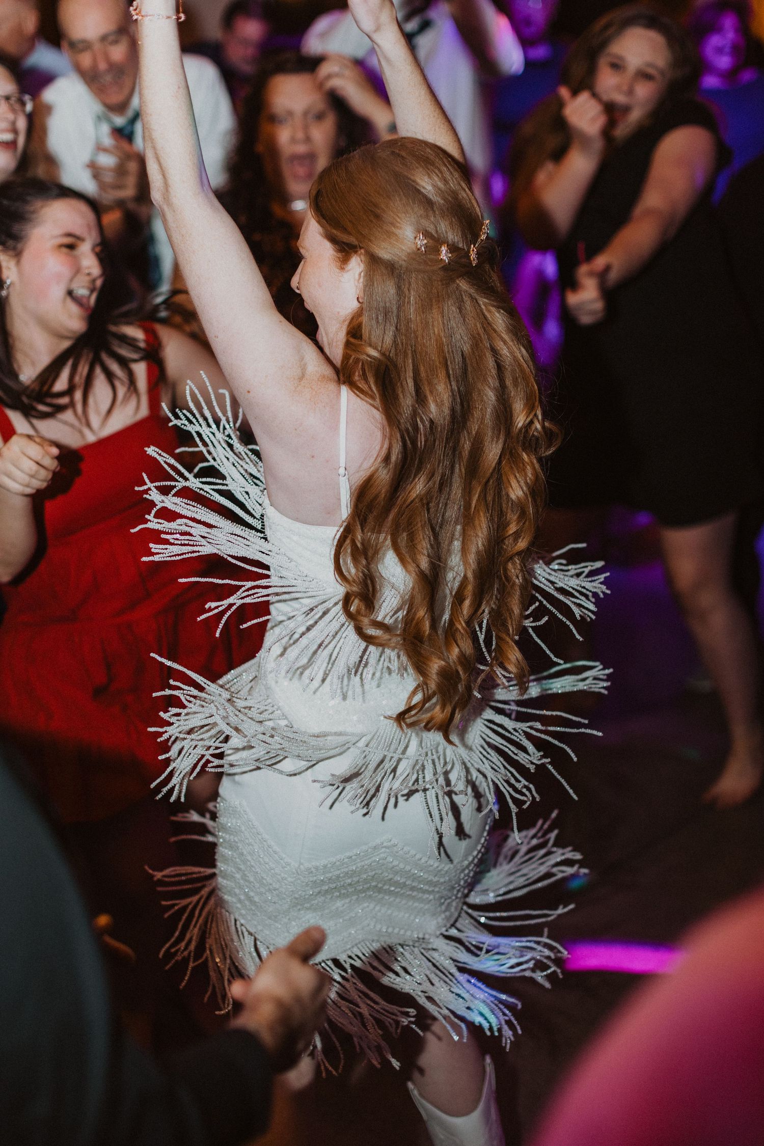 Bride in white fringe dress dancing joyfully with guests at wedding reception party.