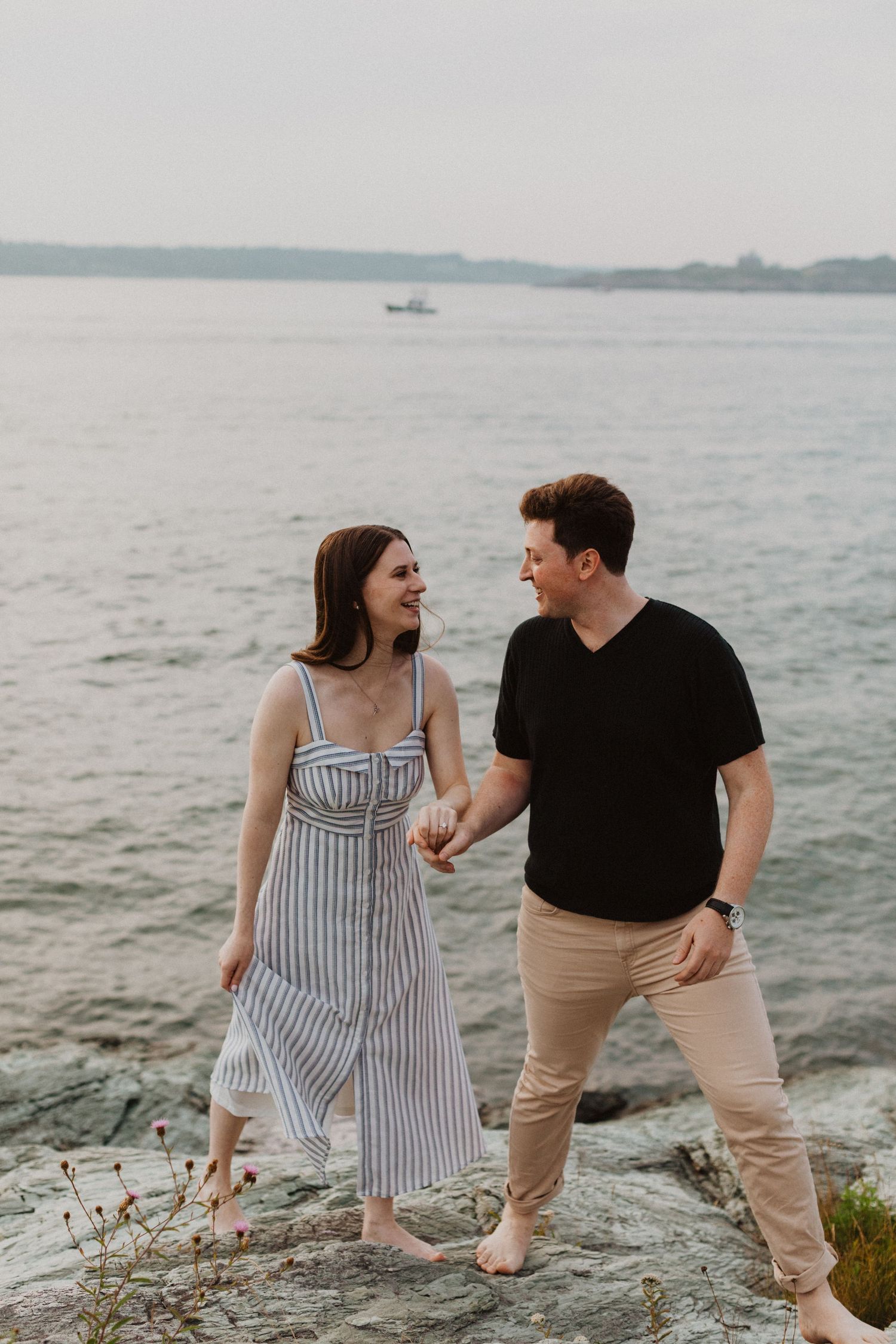 Happy couple holding hands and smiling at each other on rocky shoreline by the water.