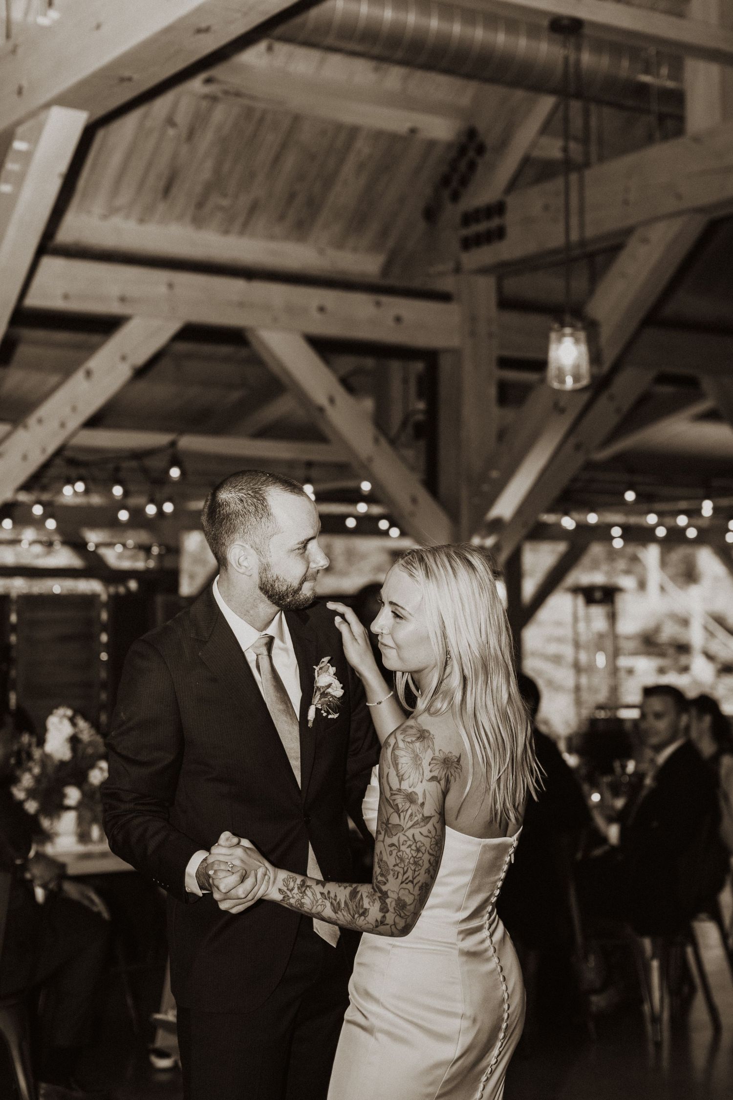 Bride and groom sharing first dance in rustic barn venue with string lights, black and white photo.