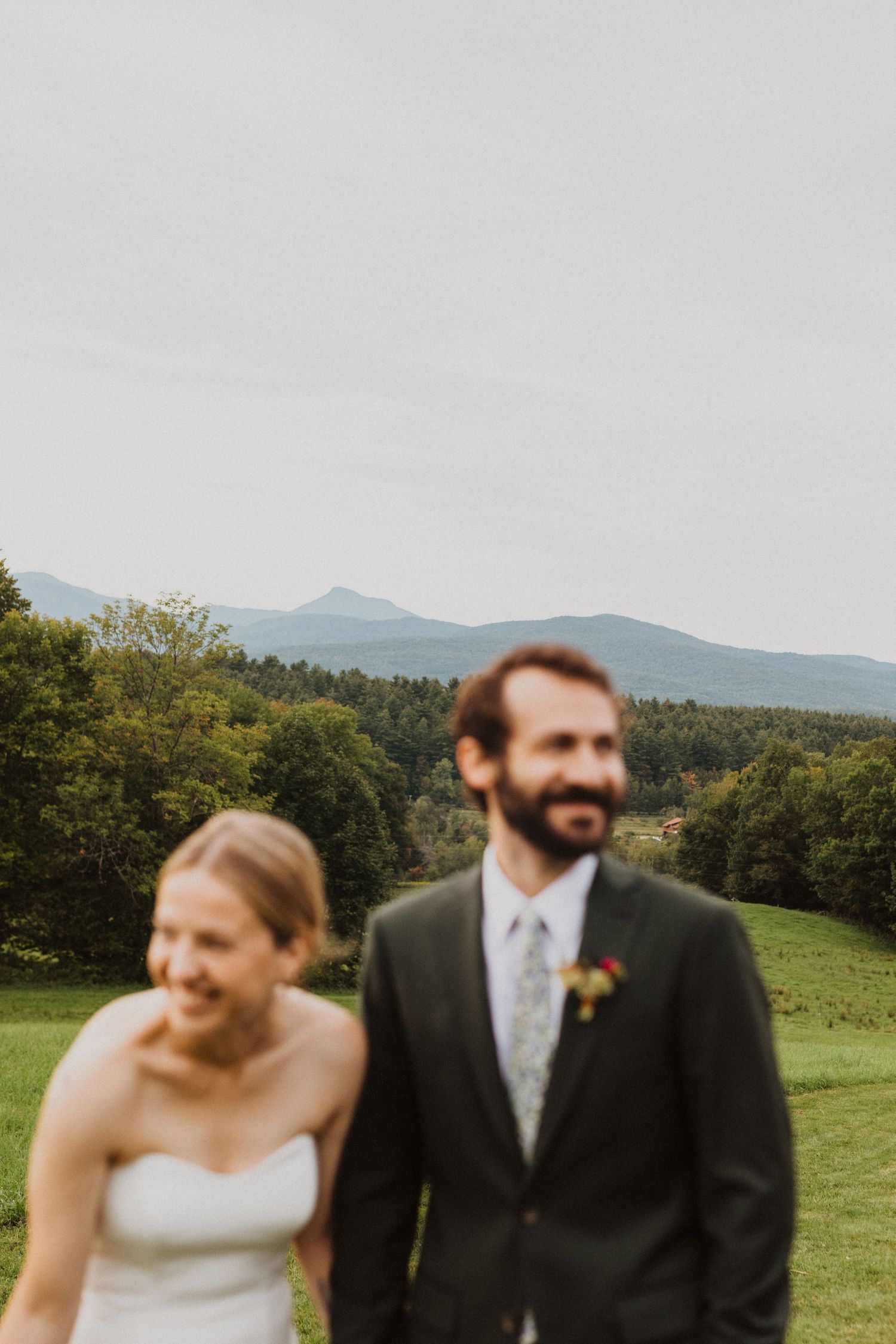 A bride and groom smile outdoors with a lush green mountain landscape visible behind them on their wedding day.