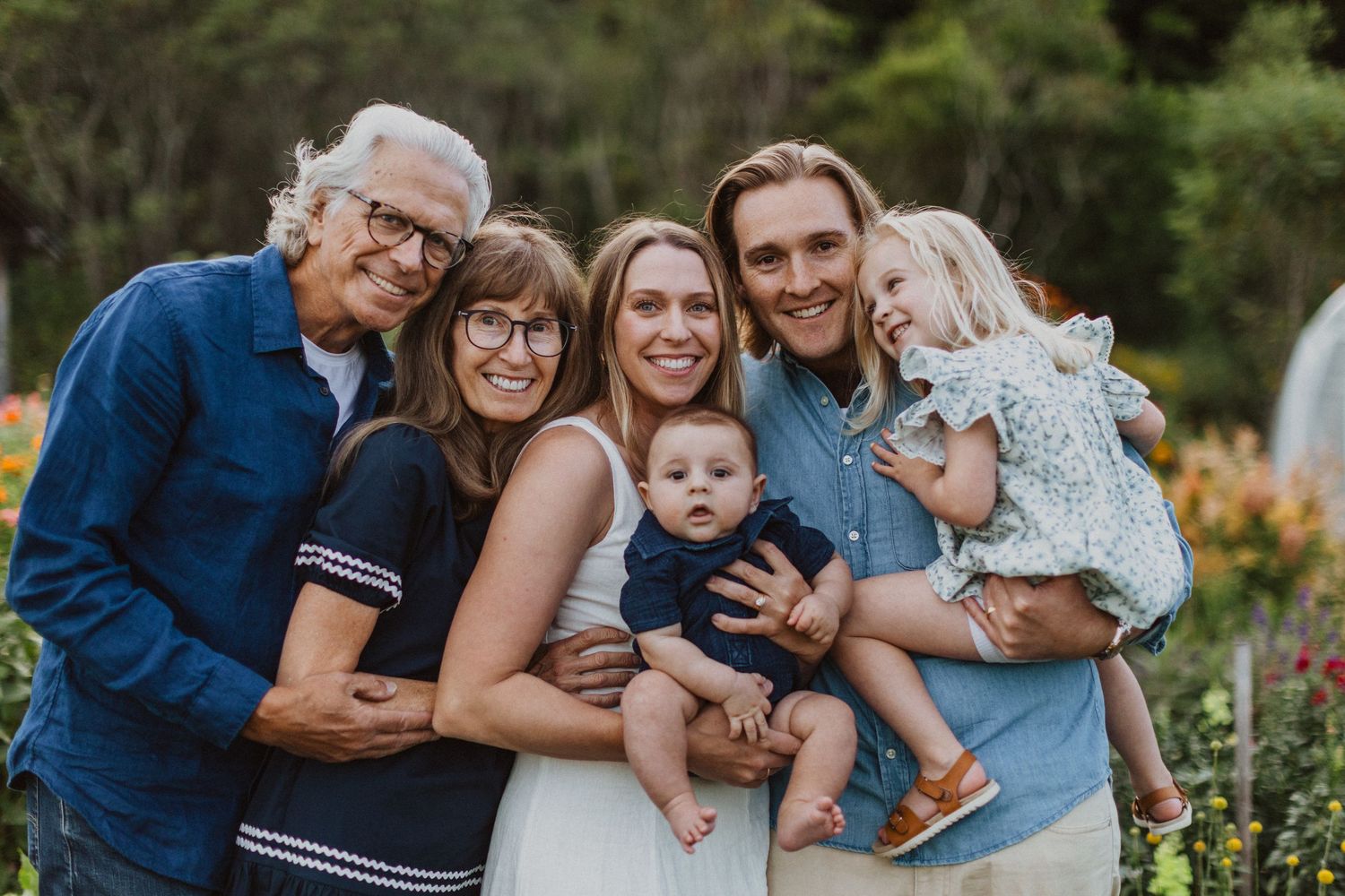 Happy family of six posing together outdoors, including grandparents, parents, baby, and toddler in a garden setting.