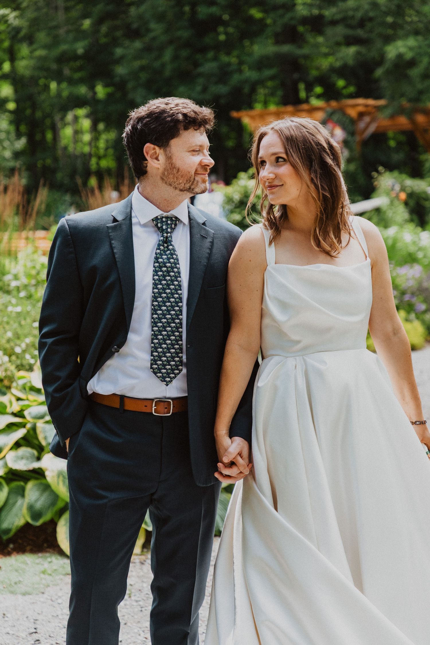 Smiling bride and groom walking hand in hand outdoors, groom in navy suit with green patterned tie, bride in white gown.
