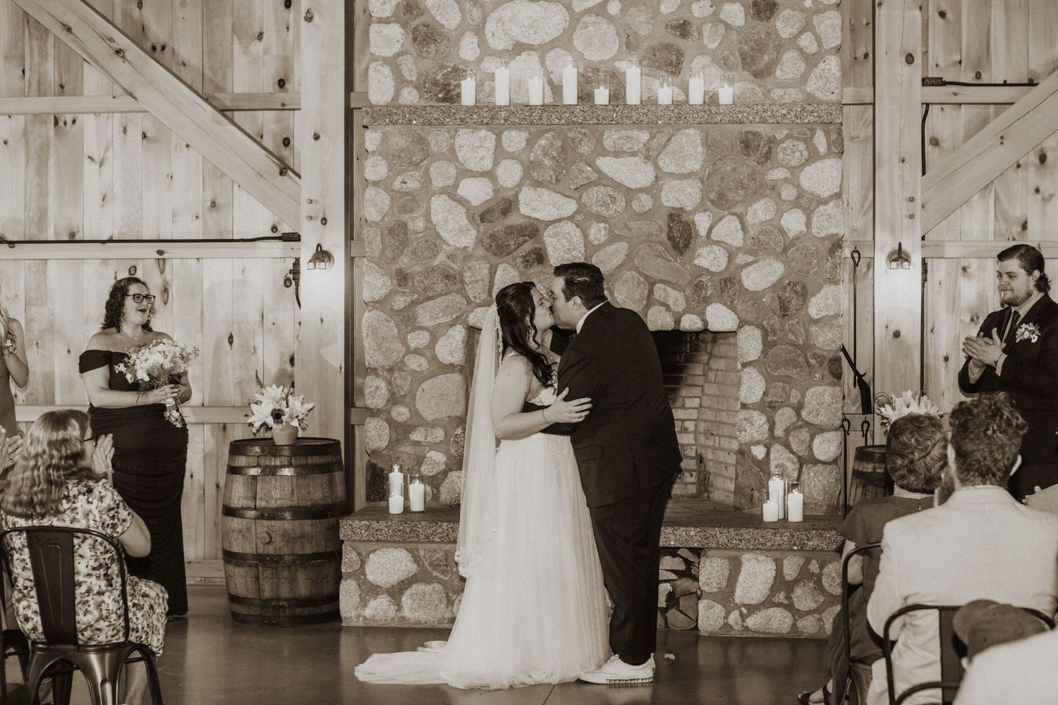 Black and white photo of bride and groom sharing first kiss at rustic barn wedding ceremony with stone fireplace.
