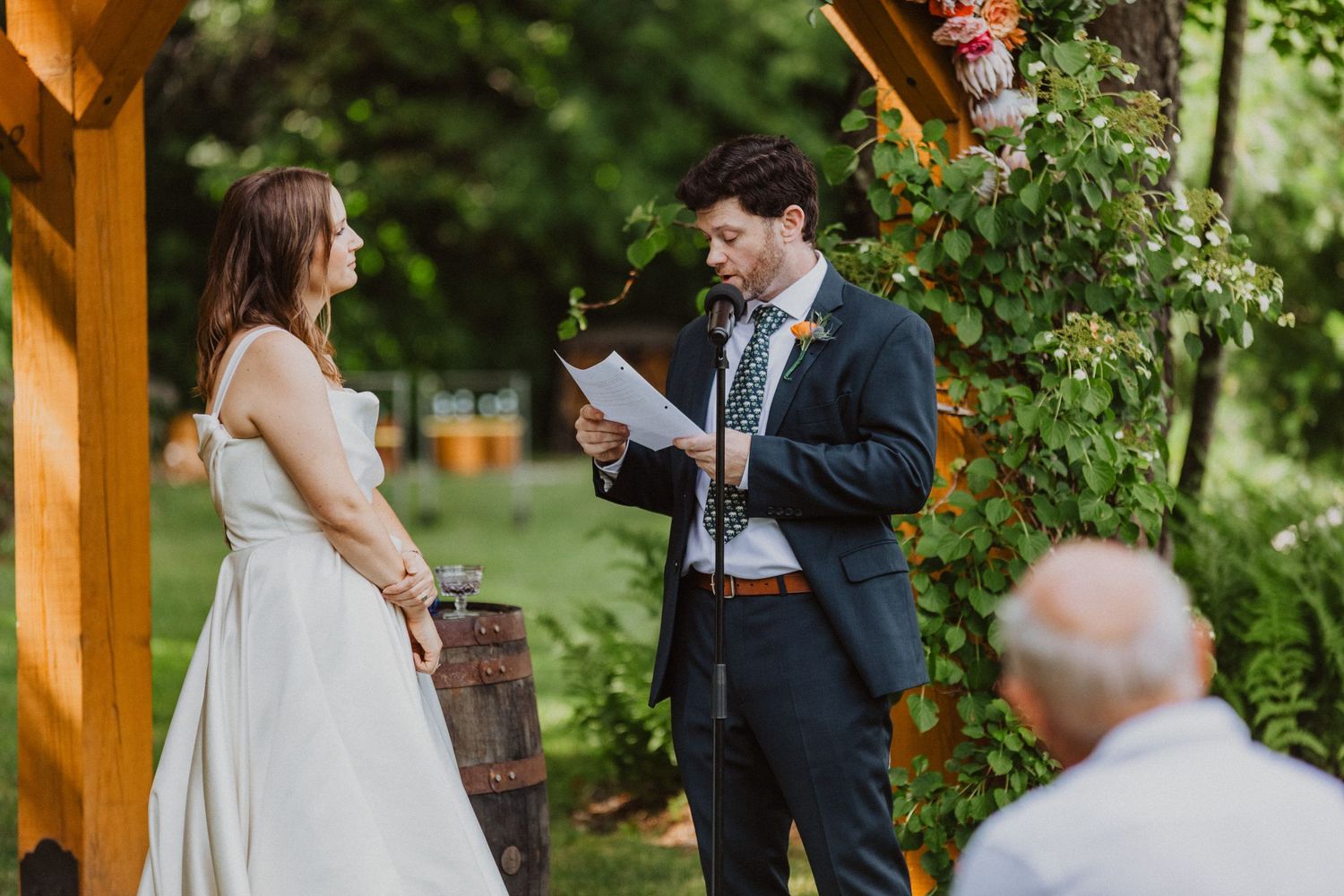 Groom reads wedding vows from paper to bride outdoors under pergola with lush greenery at rustic garden ceremony.