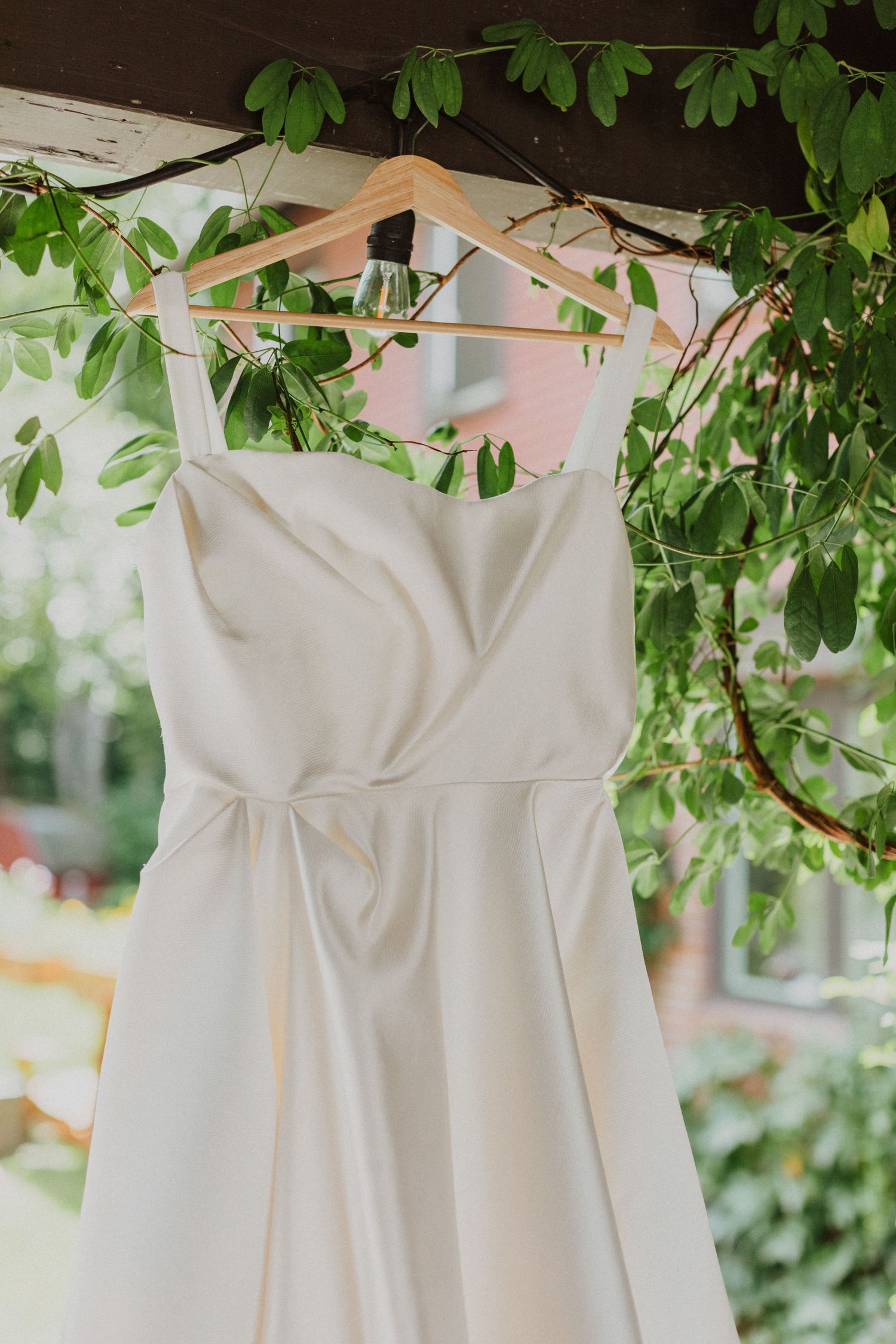 Elegant white wedding dress hanging on a wooden hanger among lush green leaves outdoors.