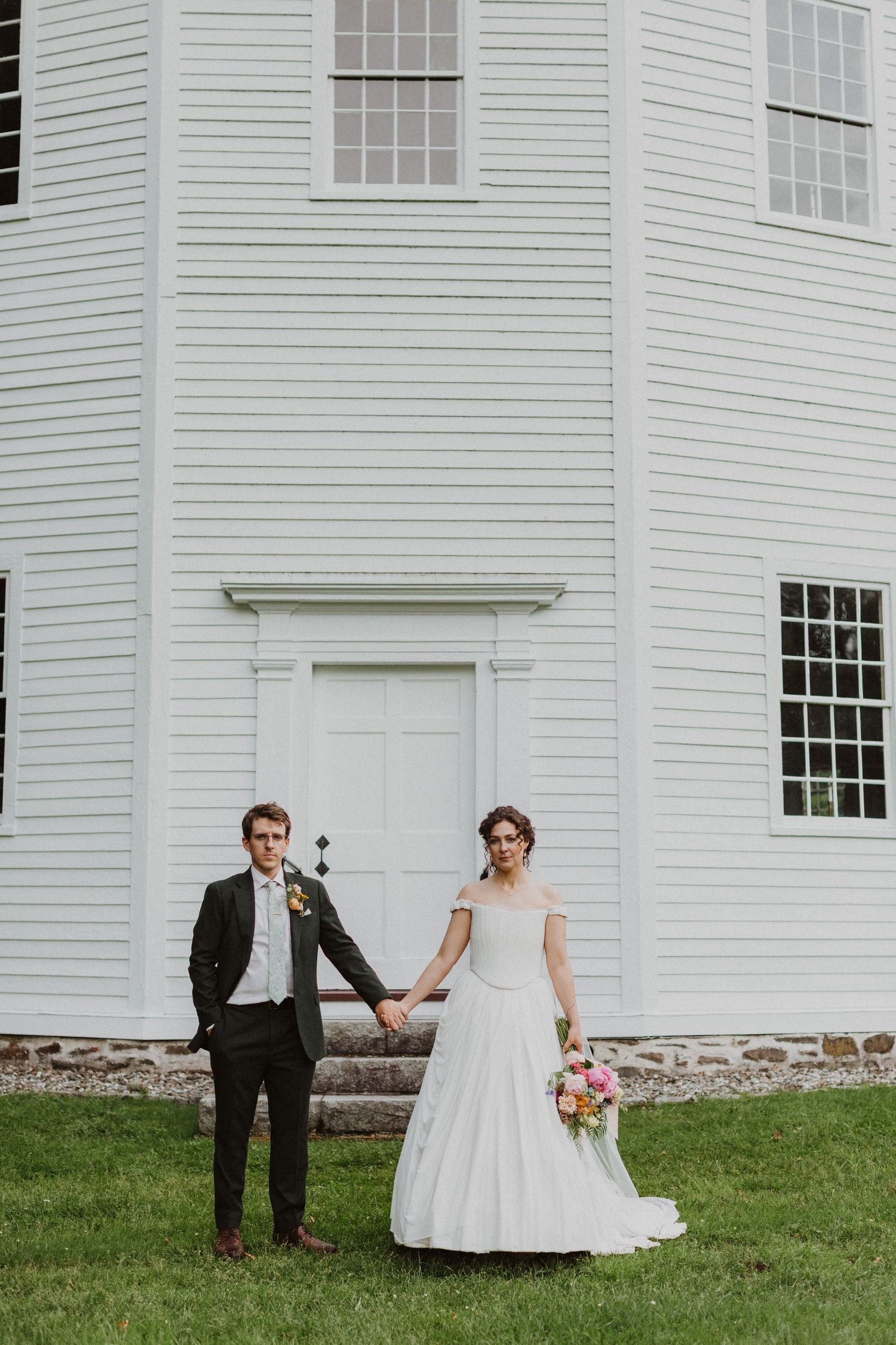 Bride and groom hold hands in front of a white colonial church on their wedding day.