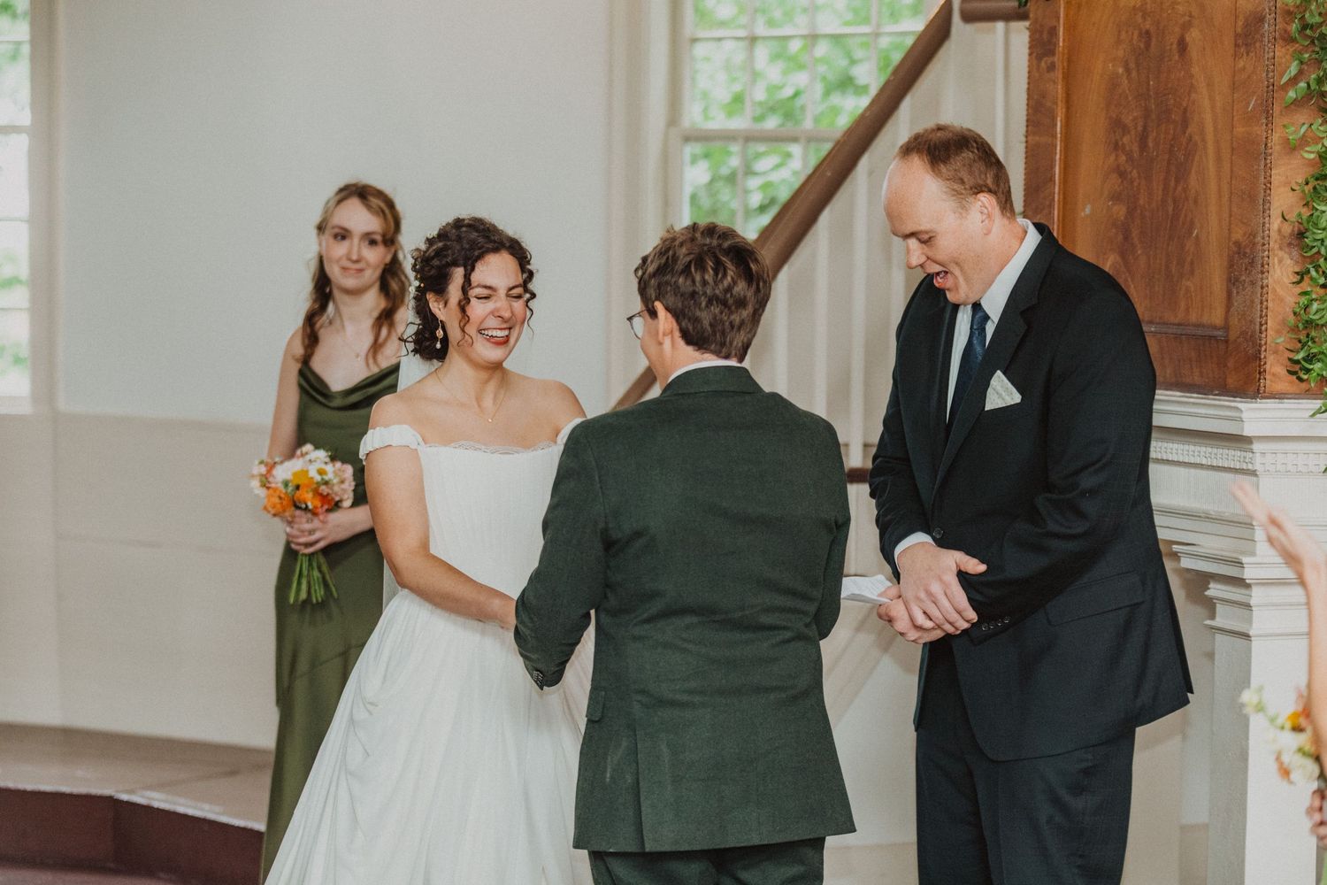 Bride smiling at groom during intimate wedding ceremony with officiant and bridesmaid in green dress.