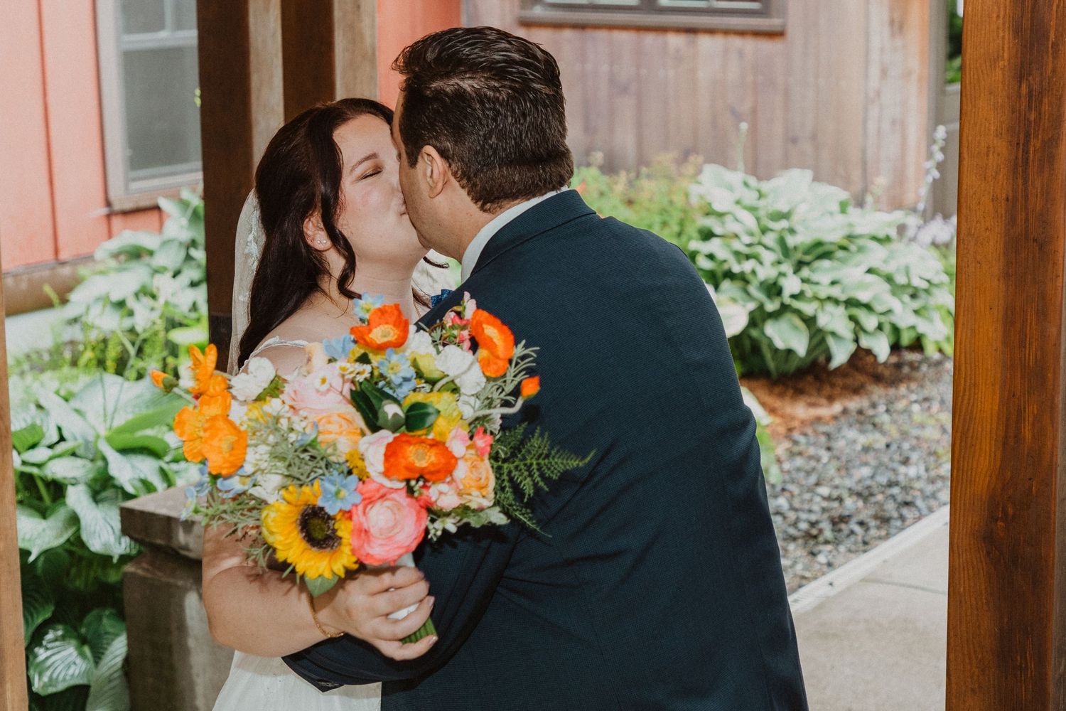 Bride and groom share a romantic kiss while bride holds a vibrant bouquet of sunflowers and orange blooms.