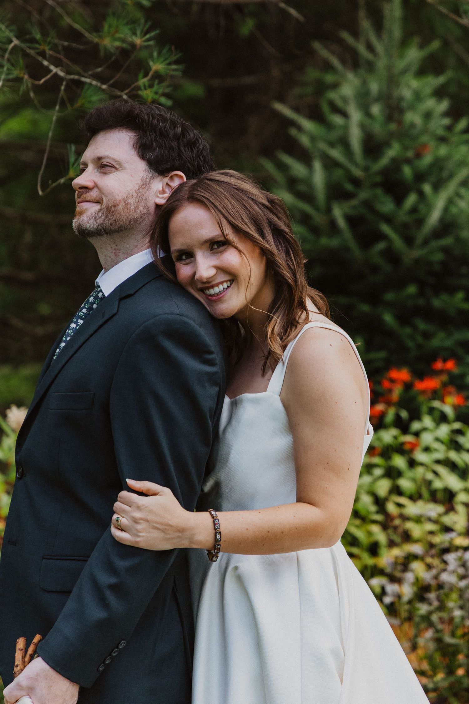 Smiling bride in white dress embraces groom in dark suit outdoors with lush green garden background.