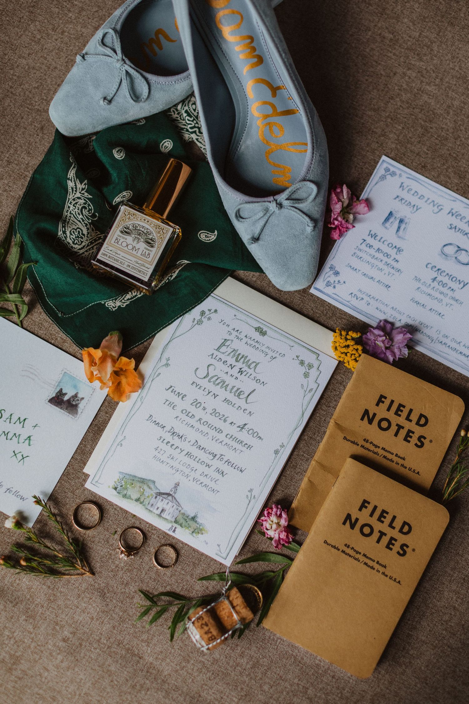 Wedding flat lay with blue suede shoes, floral invitations, Field Notes notebooks, gold rings, and perfume on neutral background.