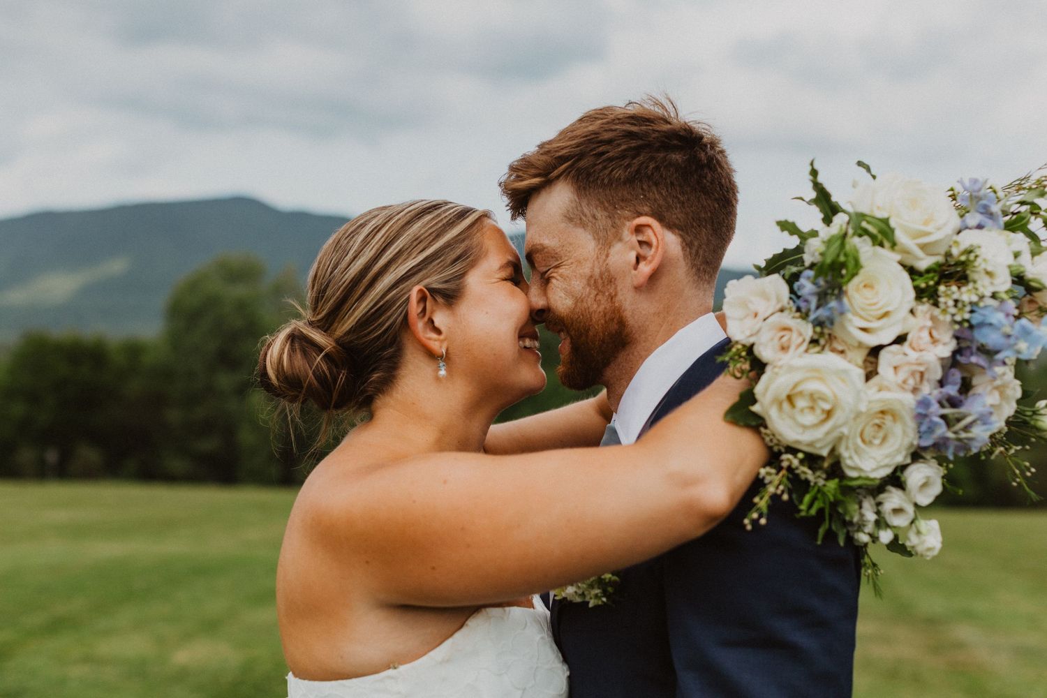 A bride holding a blue and white bouquet nearly touches foreheads with her groom against a mountain backdrop.