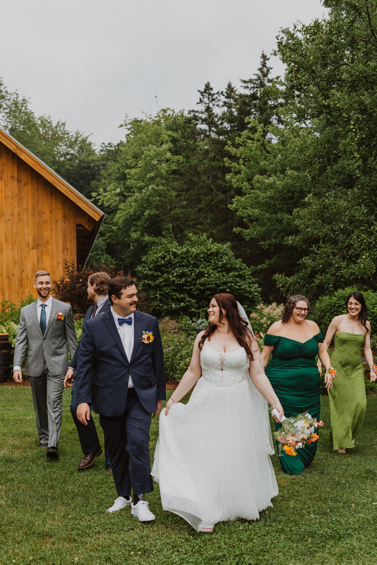 A bride and groom walk with their wedding party across a green lawn near a wooden barn surrounded by lush trees.