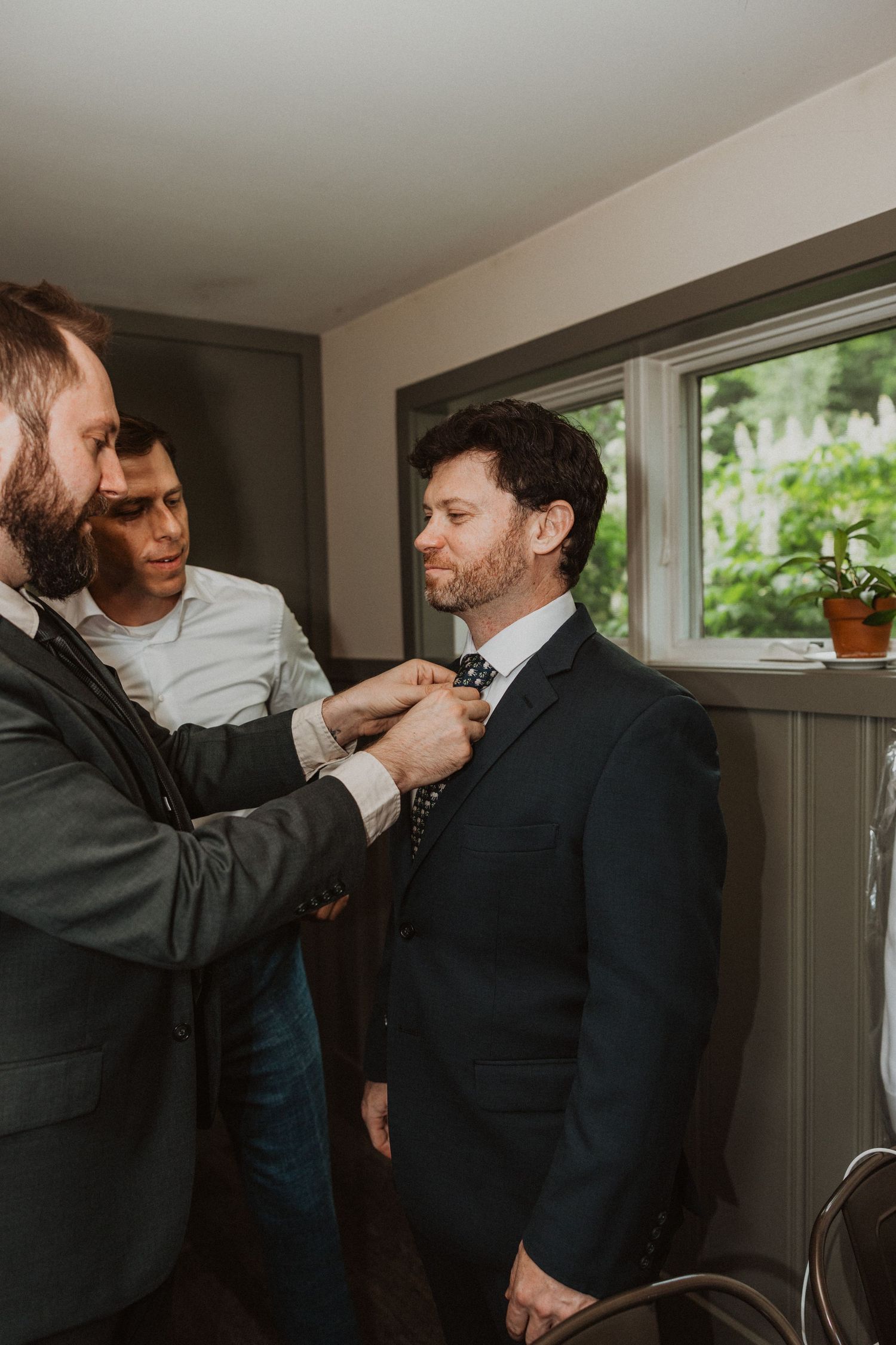 Two groomsmen helping a groom adjust his bow tie while getting ready for a wedding ceremony.