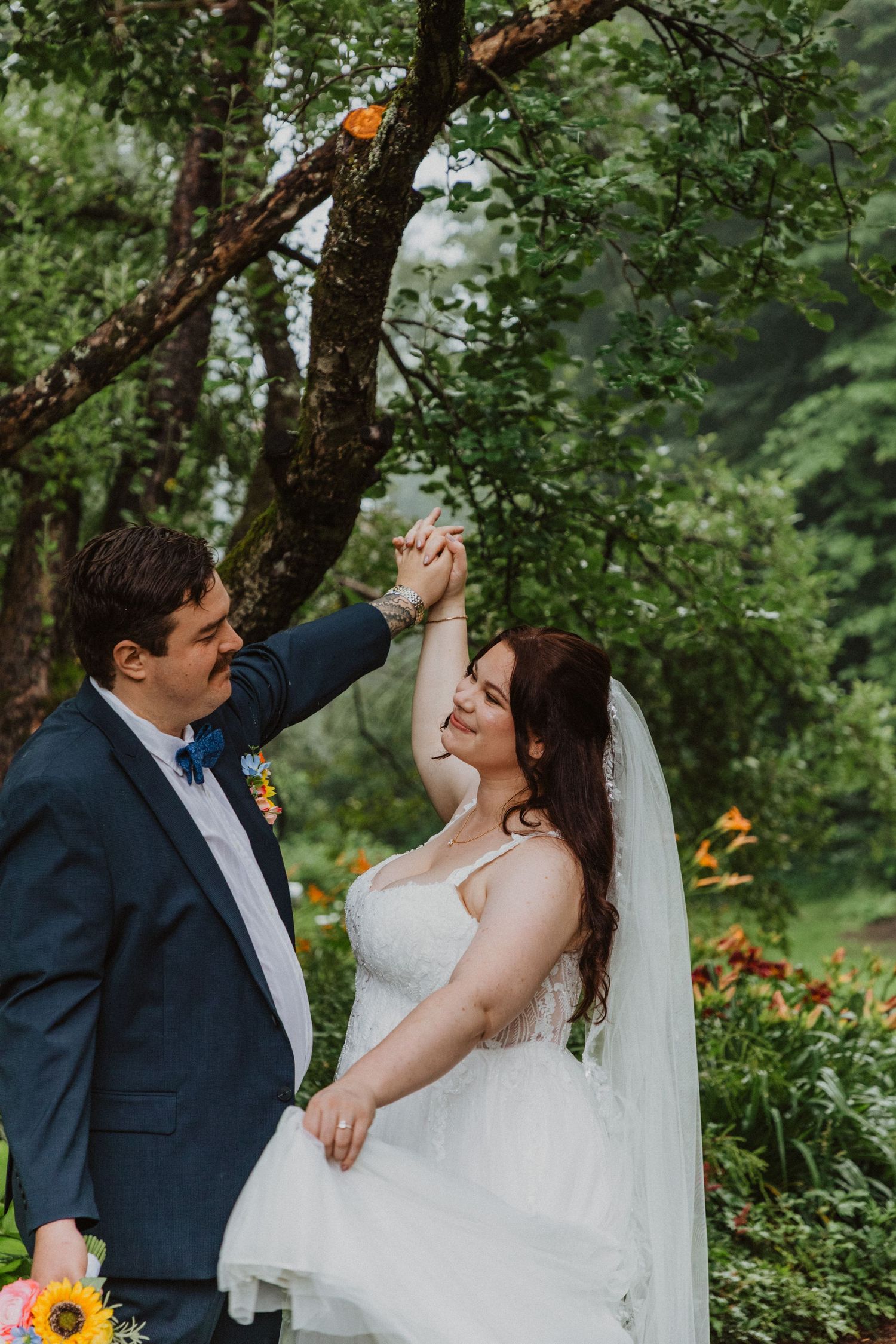 Bride and groom dancing under a tree in a lush garden, wearing a white gown and navy tuxedo with sunflowers.