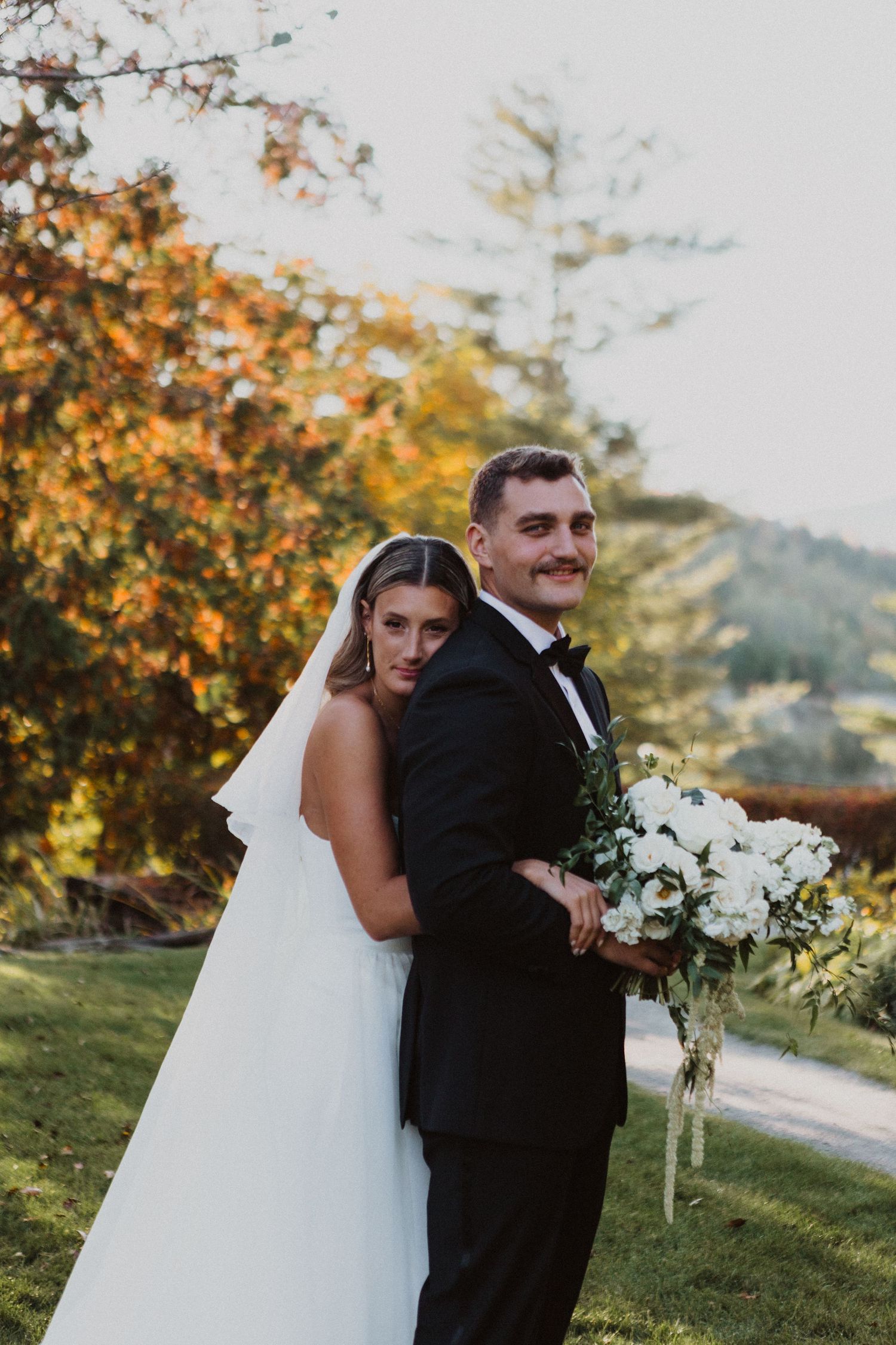 Bride leans against groom holding white bouquet as they pose together in stunning autumn scenery.