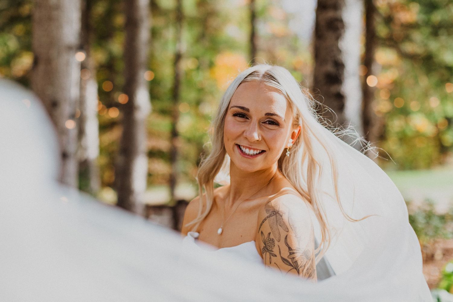 Smiling blonde bride with tattoo and flowing veil in autumn forest wedding portrait.