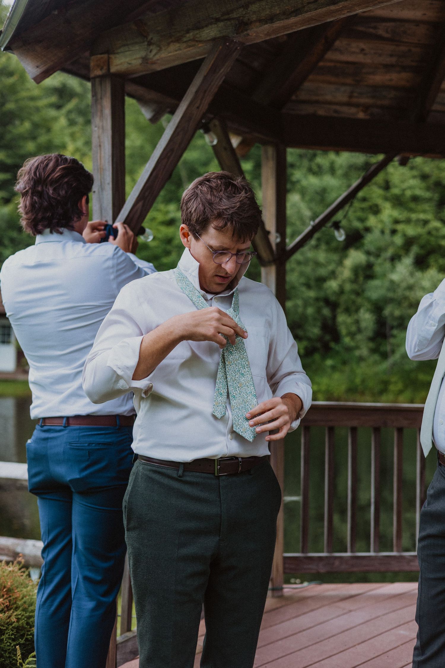 Groom adjusting his green tie while getting ready outdoors on a wooden deck before his wedding ceremony.