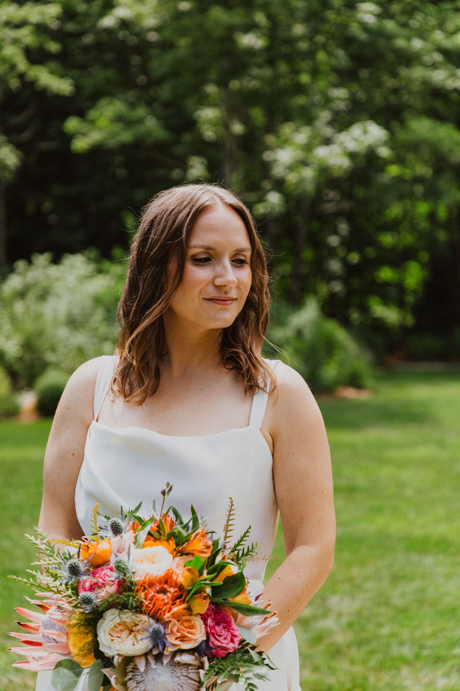 Bride in white gown holding vibrant tropical bouquet with orange protea and wildflowers in lush garden.