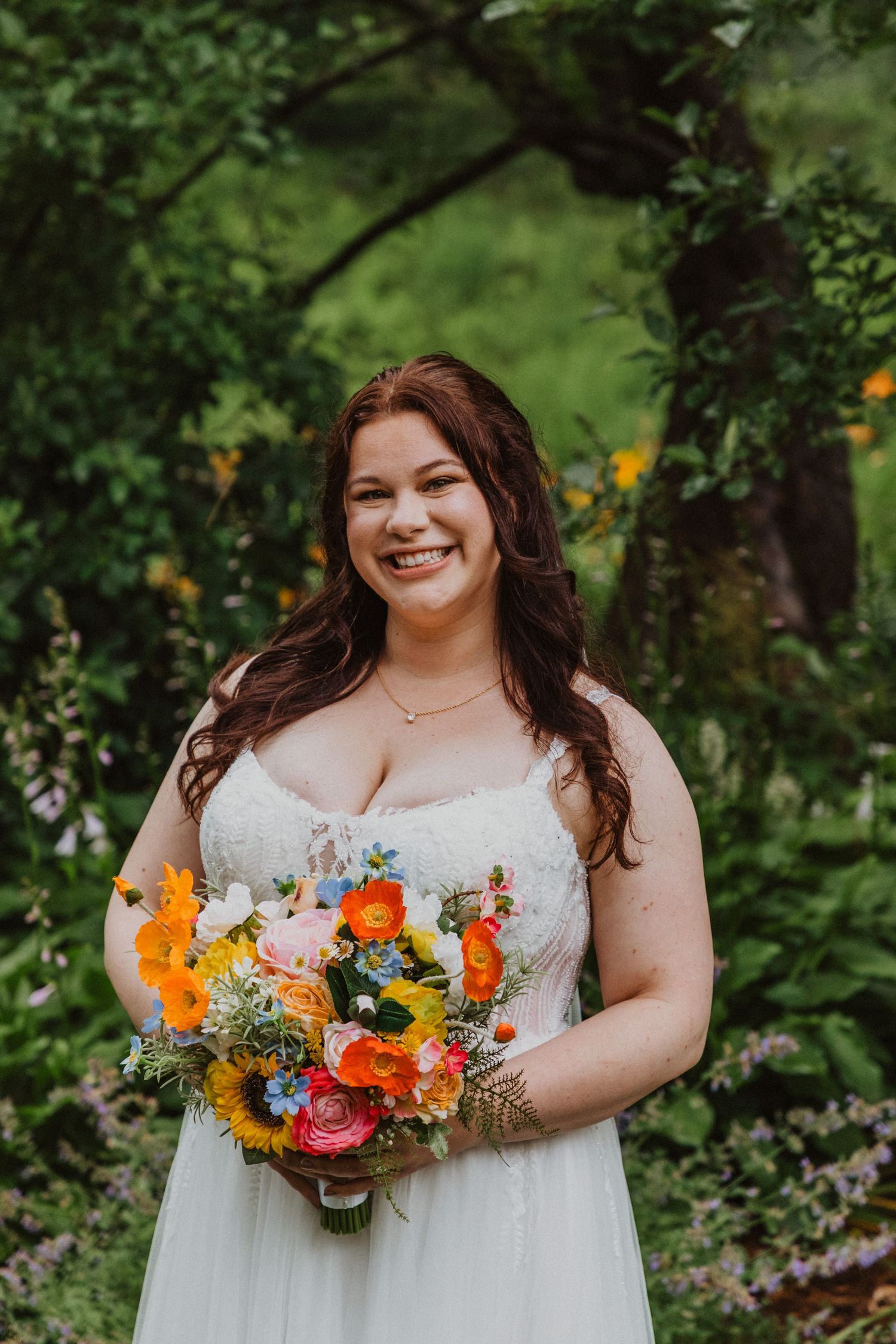 Smiling bride in white gown holds colorful bouquet of orange, yellow, and blue flowers outdoors in lush green garden.