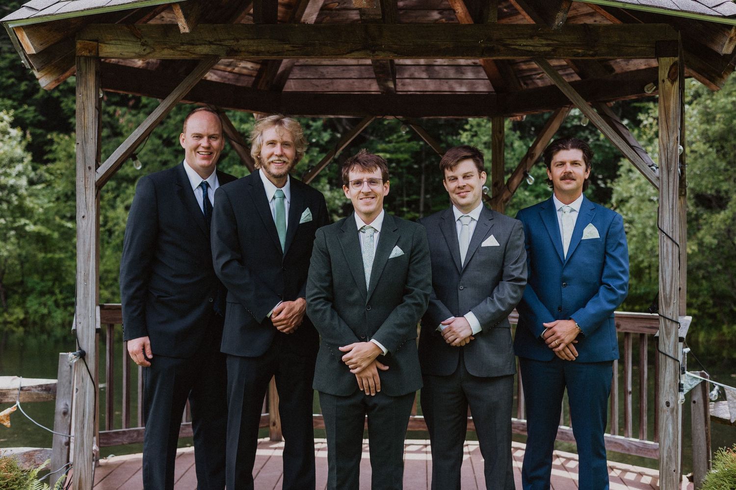 Five men in suits posing together under a wooden gazebo at an outdoor wedding venue surrounded by trees.